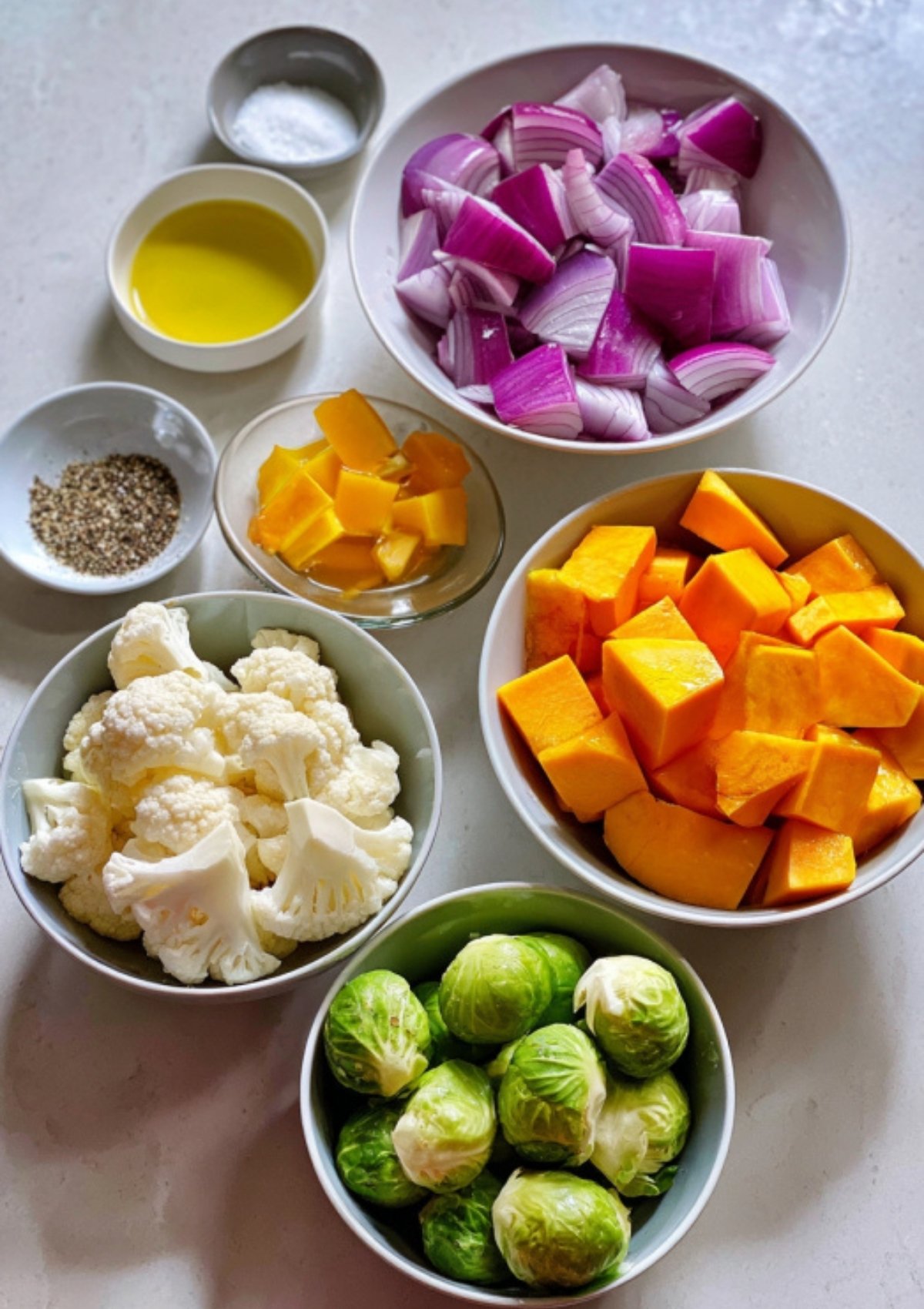 Bowls filled with chopped red onion, butternut squash, cauliflower, brussels sprouts, olive oil, salt, and pepper arranged on a white countertop.