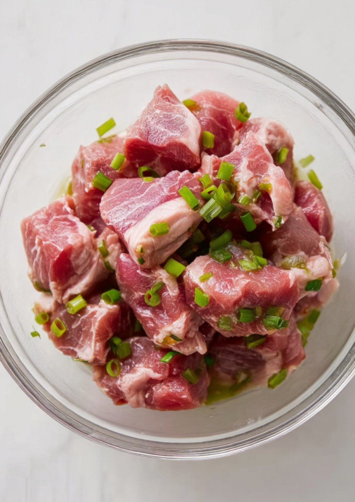 Chunks of raw pork marinating with chopped green onions in a glass bowl.