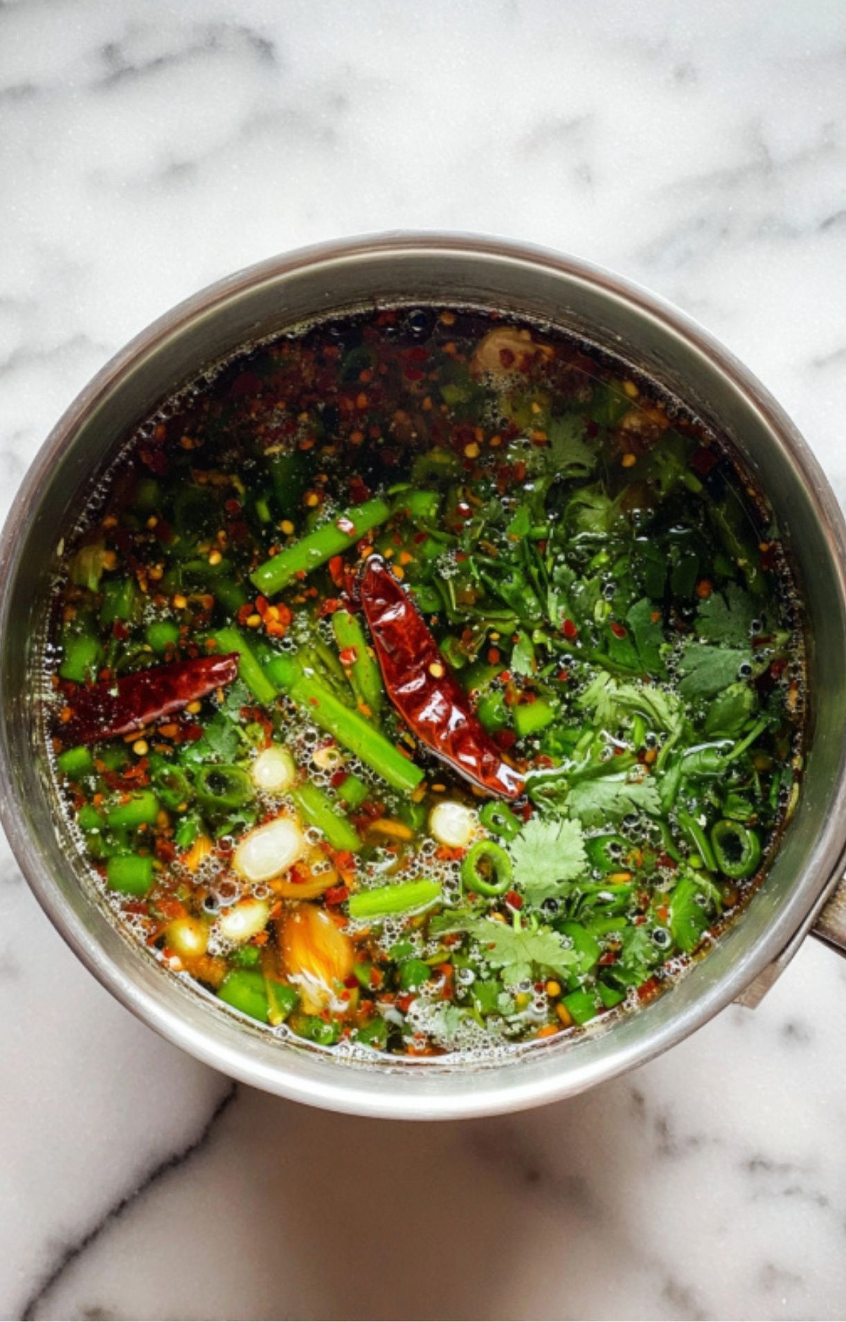Pot of hot oil infusing with cilantro, green onions, dried chilies, and spices for a bold Asian chili sauce.