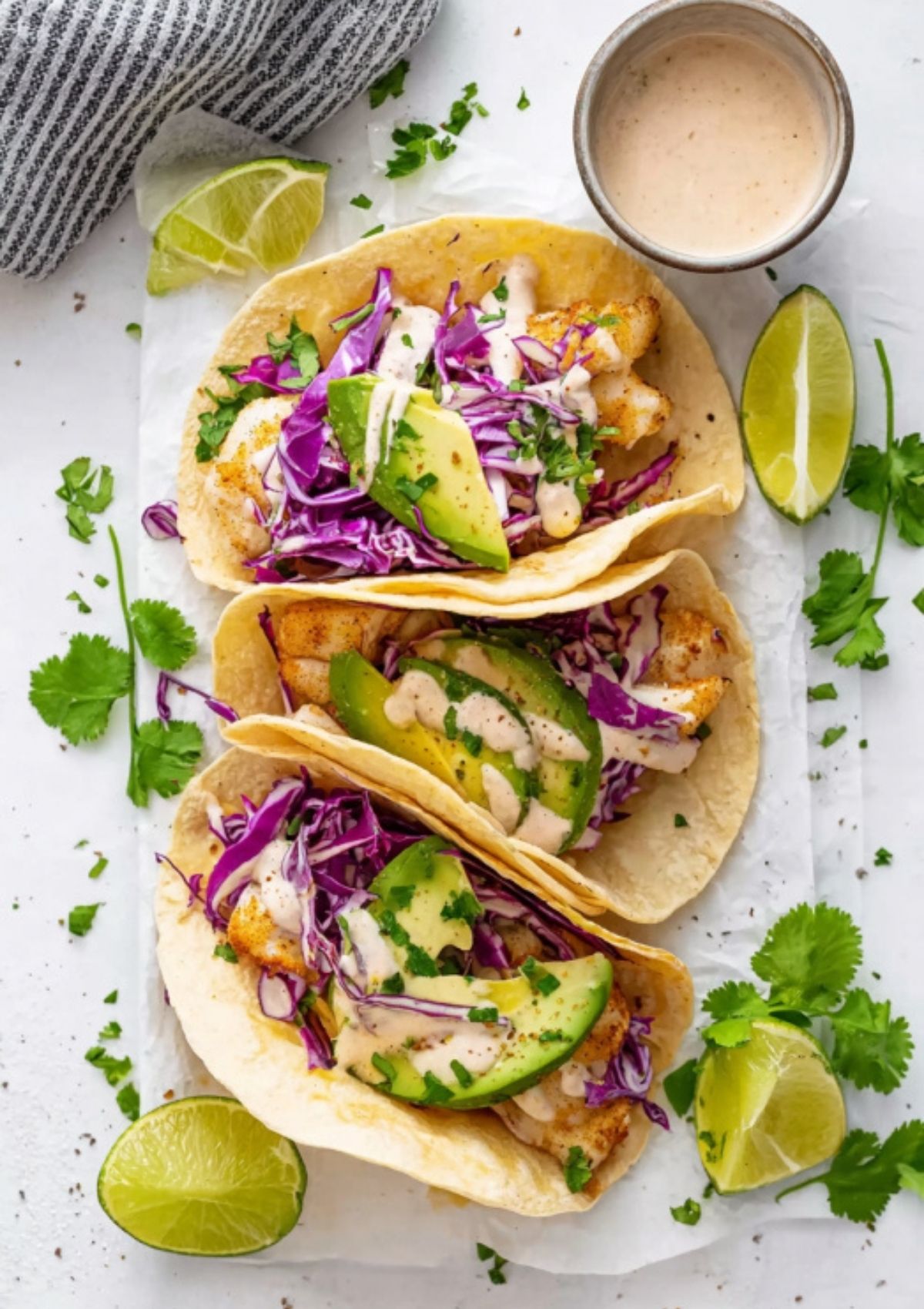 Overhead view of crispy fish tacos in tortillas, loaded with shredded red cabbage, avocado, cilantro, and creamy sauce, with limes and cilantro on the side.
