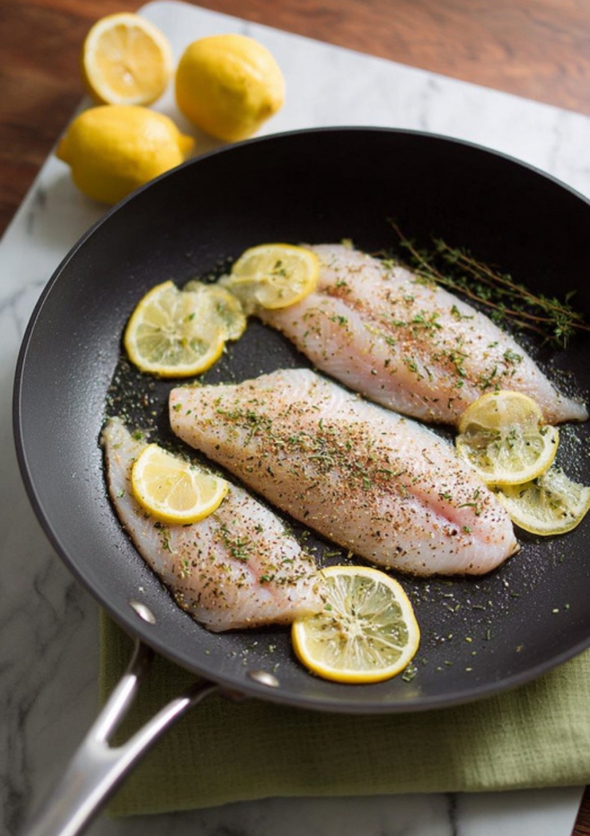 Skillet with tilapia fillets, lemon slices, and sprinkled herbs, ready to bake for lemon herb tilapia.
