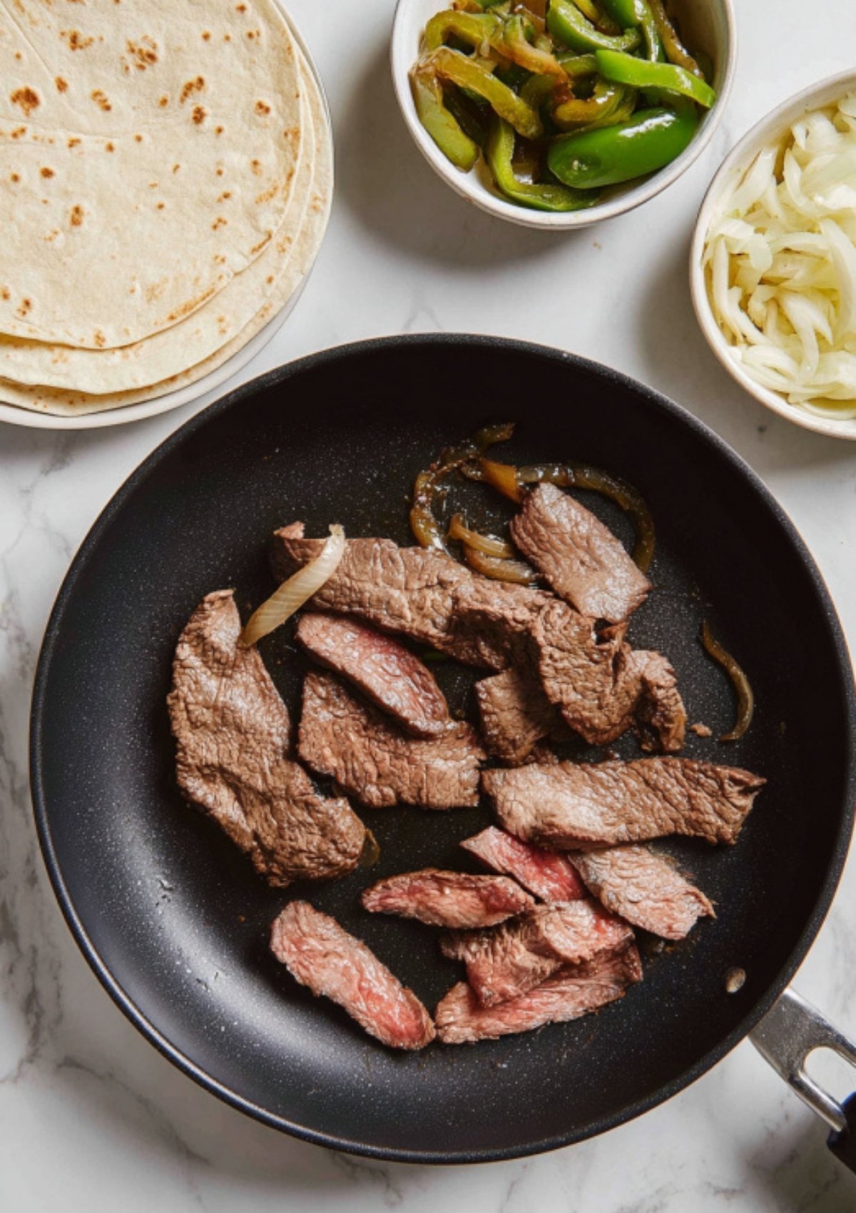 Strips of steak cooking in a skillet, with bowls of sautéed peppers, onions, and a stack of tortillas on the counter.