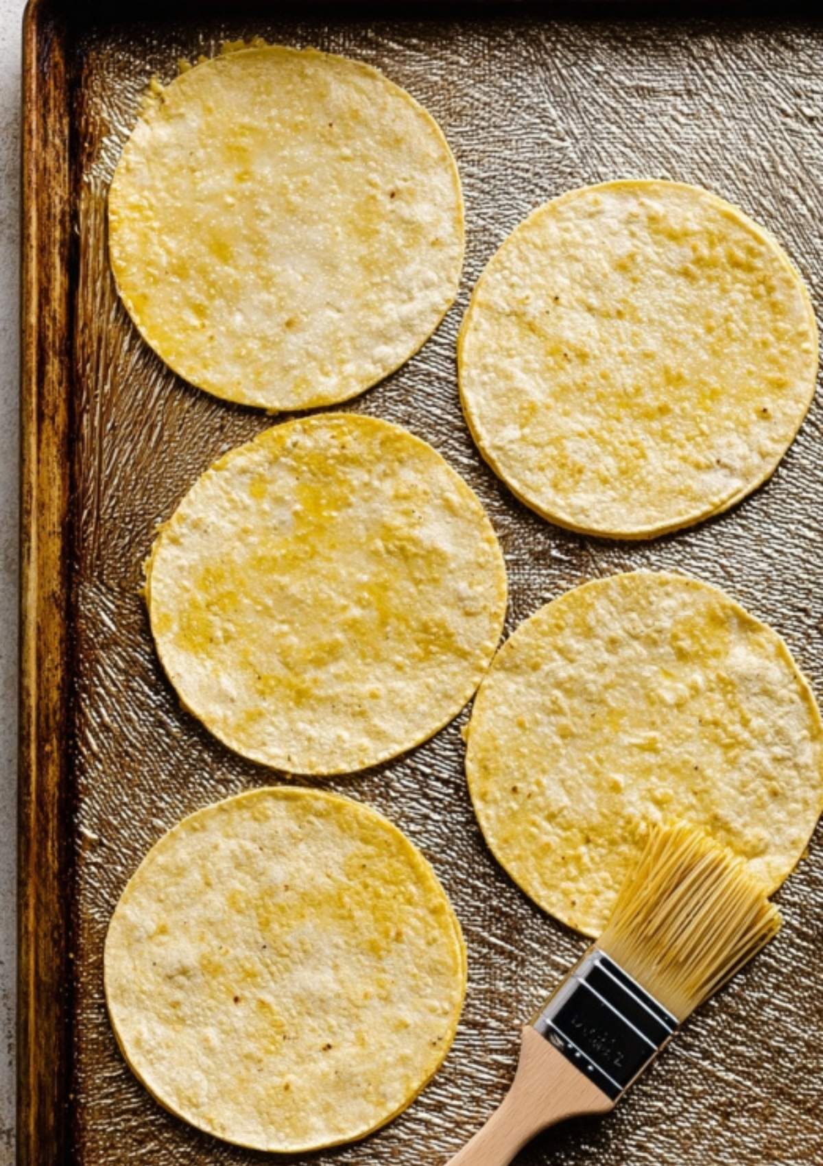 Corn tortillas brushed with oil on a baking sheet with a pastry brush, ready for filling and baking.