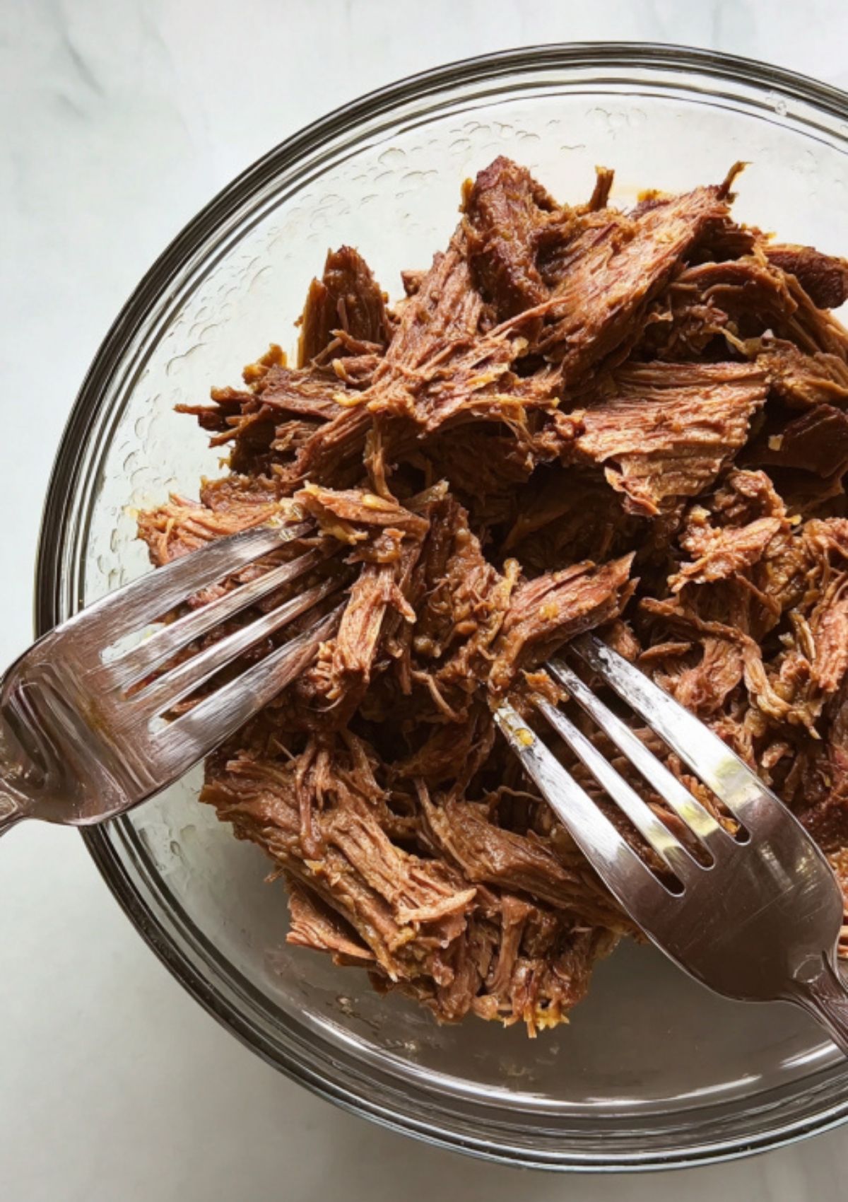 Juicy shredded Mississippi pot roast pulled apart with two forks in a glass bowl, ready for sandwiches or serving over potatoes.