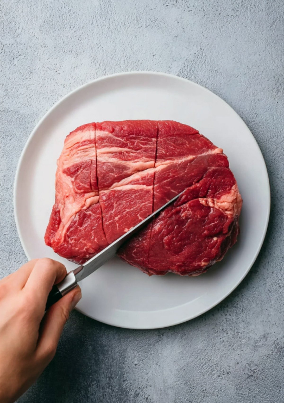 Hand using a knife to score raw chuck roast on a white plate—step for tenderizing and adding deep flavor.