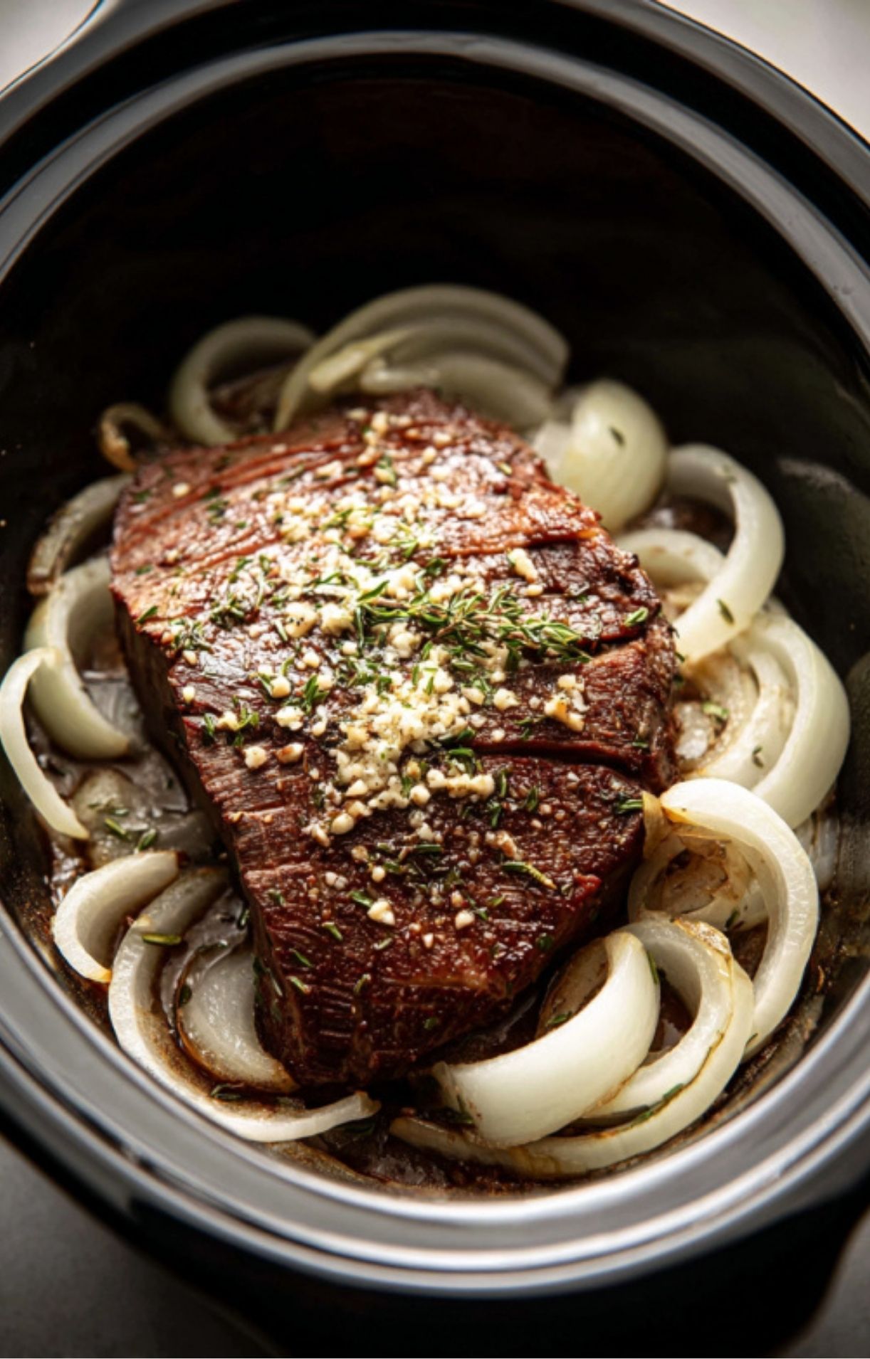 Tender beef chuck roast in a crockpot surrounded by sliced onions, seasoned with garlic and herbs, perfect for a hearty family meal.
