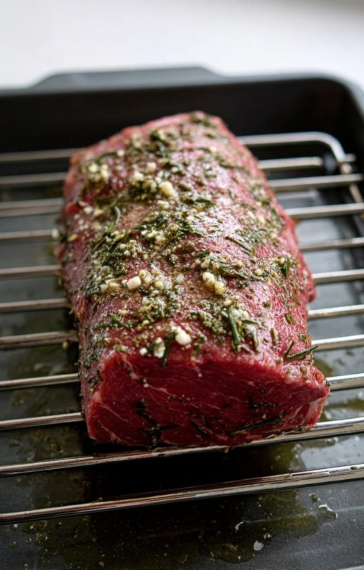 Raw beef roast coated with chopped herbs and garlic, placed on a rack before roasting