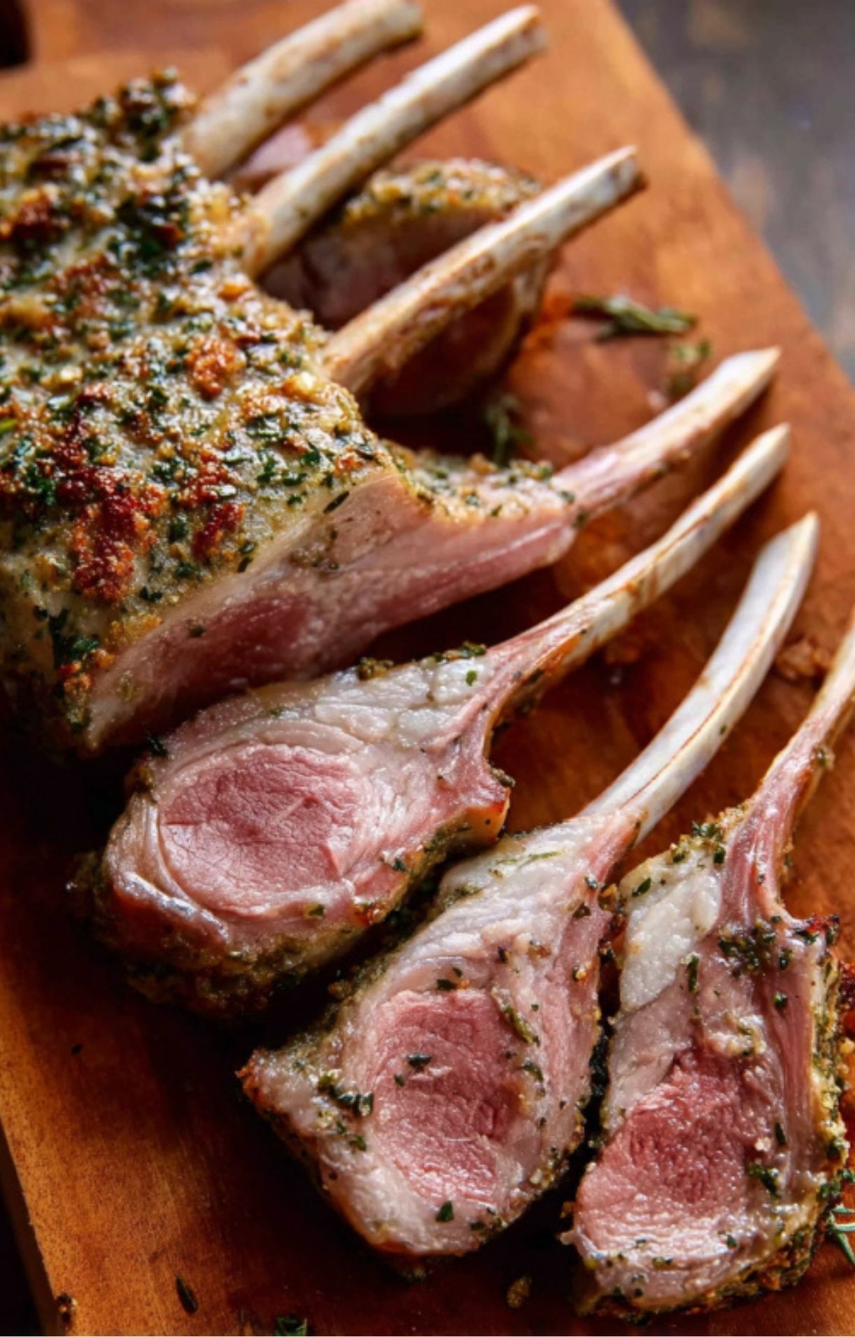 Freshly sliced rack of lamb coated in a roasted garlic and herb crust, displayed on a wooden board.