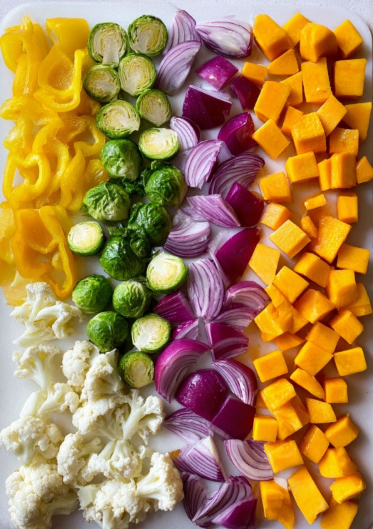 Brightly colored vegetables—yellow bell pepper, brussels sprouts, red onion, cauliflower, and butternut squash—lined up on a sheet pan, ready for roasting.