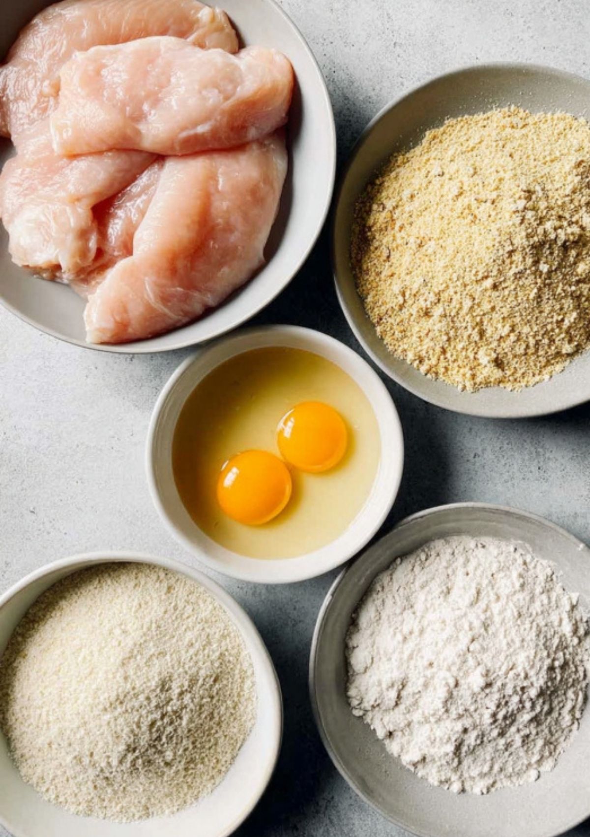 Bowls of raw chicken, eggs, flour, breadcrumbs, and parmesan cheese—ingredients for chicken parmesan sandwiches.