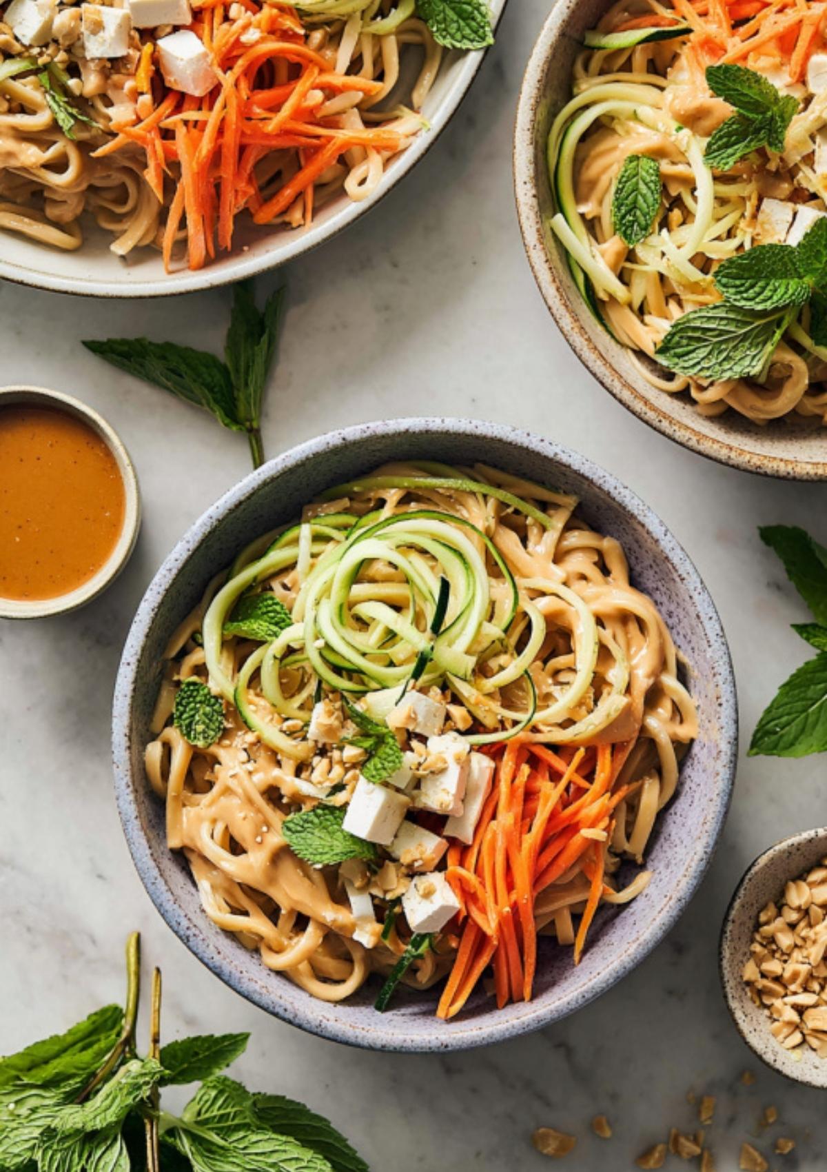 Bowls of Thai peanut noodles topped with tofu, shredded carrots, cucumber, mint leaves, and crushed peanuts.