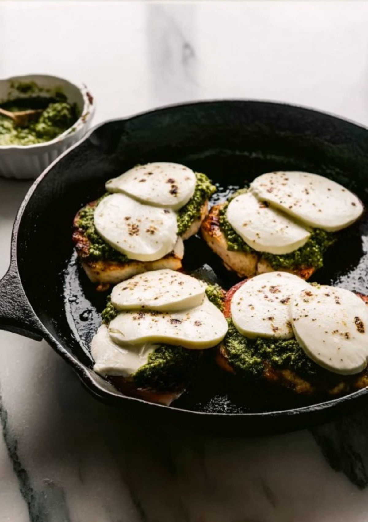 Chicken breasts in a skillet, topped with basil pesto and thick mozzarella slices for Caprese chicken.