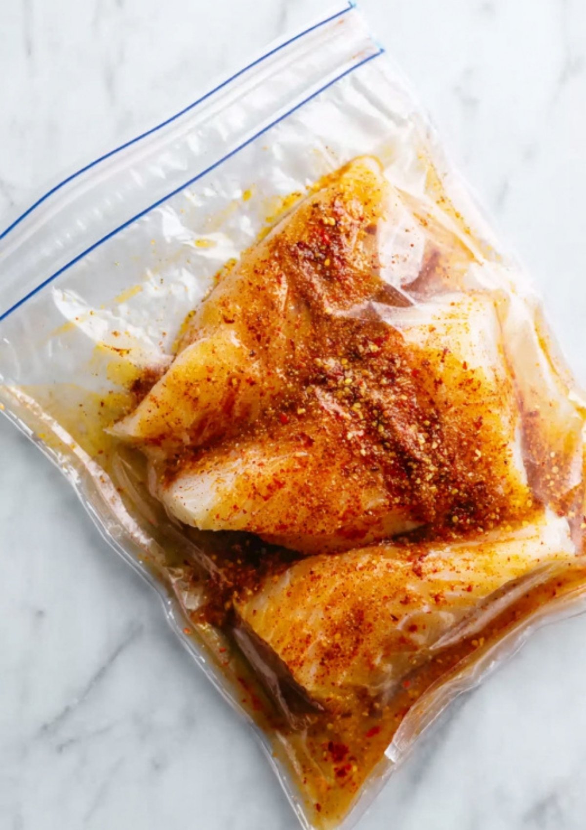 White fish fillets marinating in olive oil, chili powder, and spices inside a zip-top bag, step for juicy, flavorful crispy fish tacos.