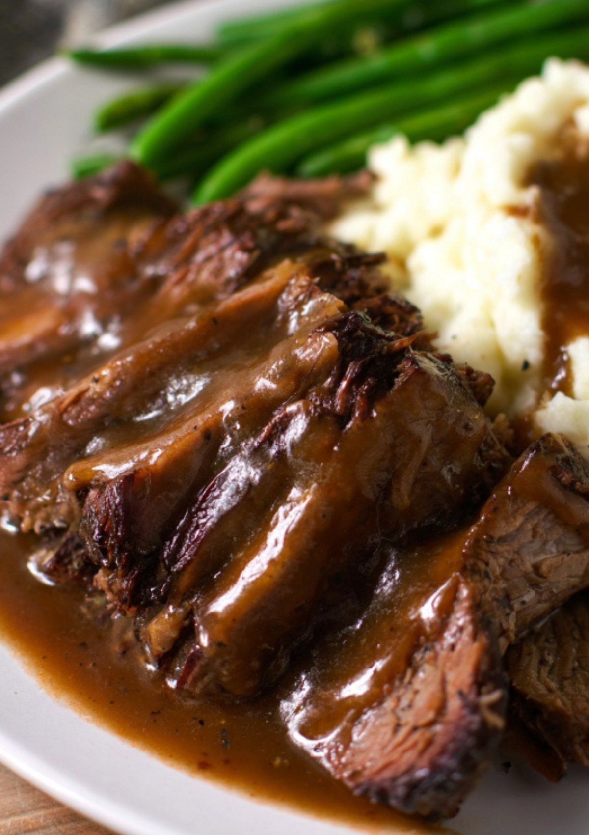 Slices of slow-cooked chuck roast covered in homemade gravy, served with mashed potatoes and green beans, classic comfort food