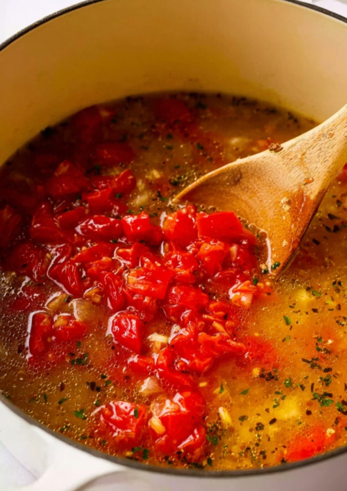 Chopped tomatoes and broth in a pot being stirred with a wooden spoon for creamy tomato tortellini soup.