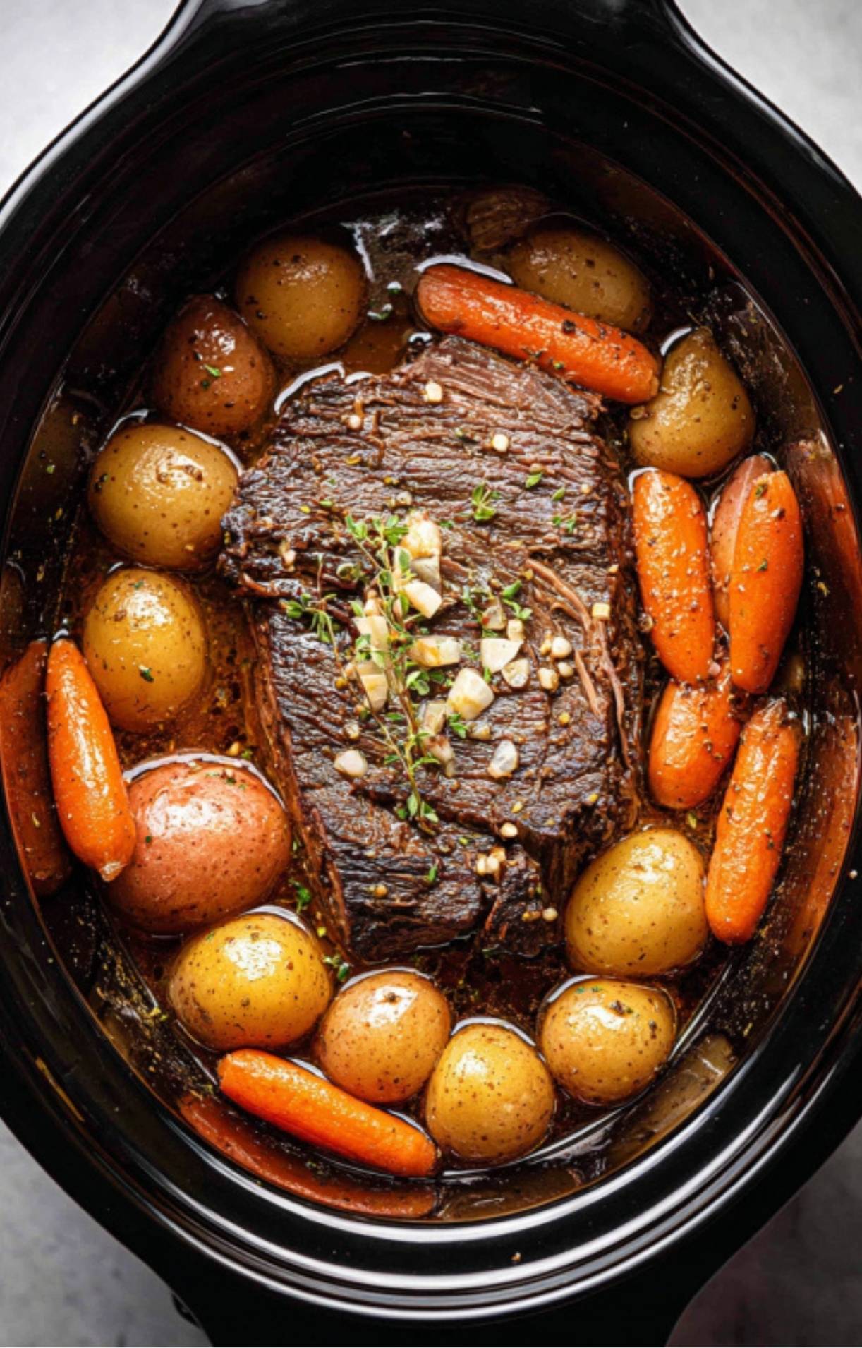 Chuck roast, baby potatoes, and carrots simmering together in a slow cooker, seasoned with herbs and garlic for a delicious dinner.