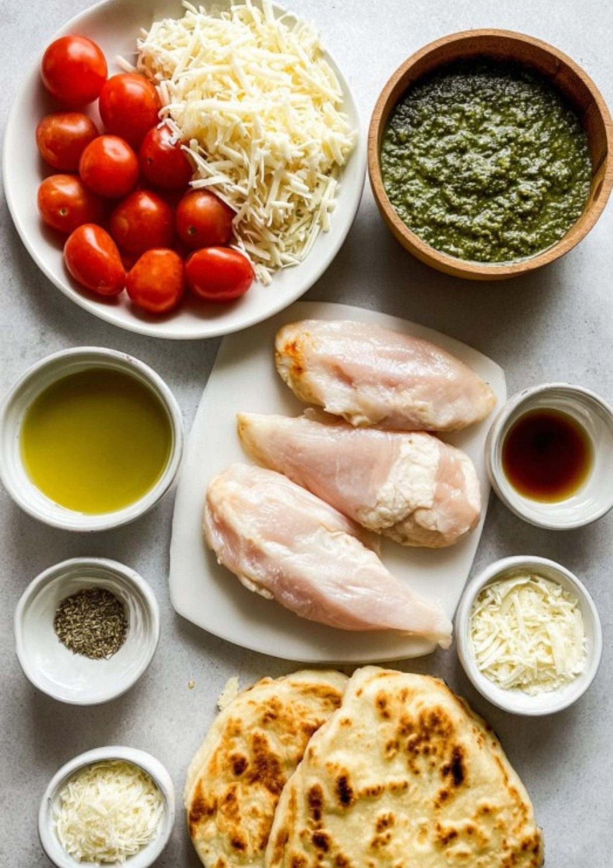 Ingredients for pesto chicken flatbread on a white surface: chicken, cheese, tomatoes, pesto, flatbread, olive oil, and spices.