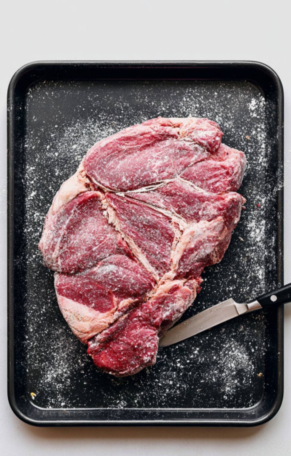 Chuck roast coated in flour on a tray with a knife, prepped for searing