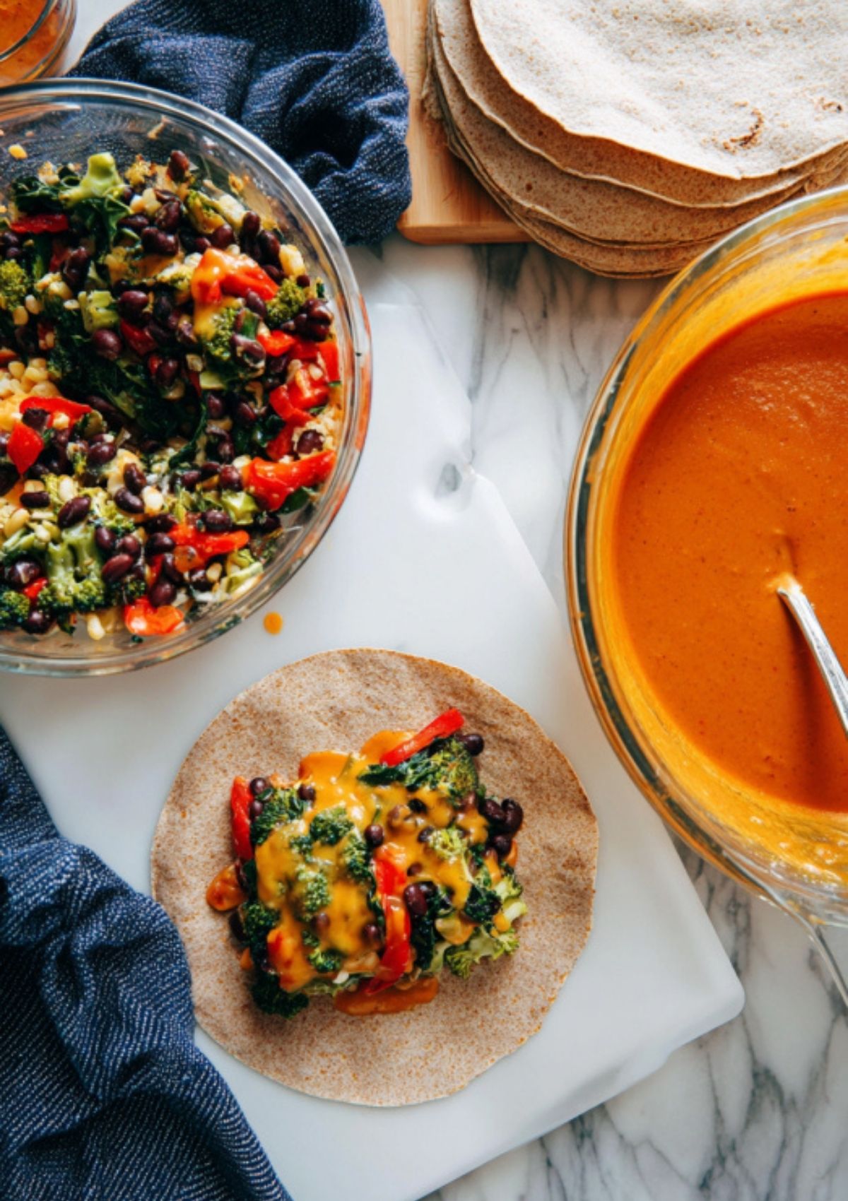 Tortilla topped with black bean, broccoli, and red pepper filling, next to bowls of enchilada sauce and extra tortillas, step-by-step for enchiladas.