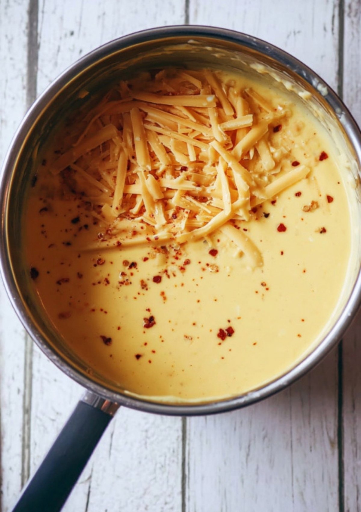 Saucepan filled with freshly made cheddar cheese sauce, sprinkled with shredded cheese and red pepper flakes.