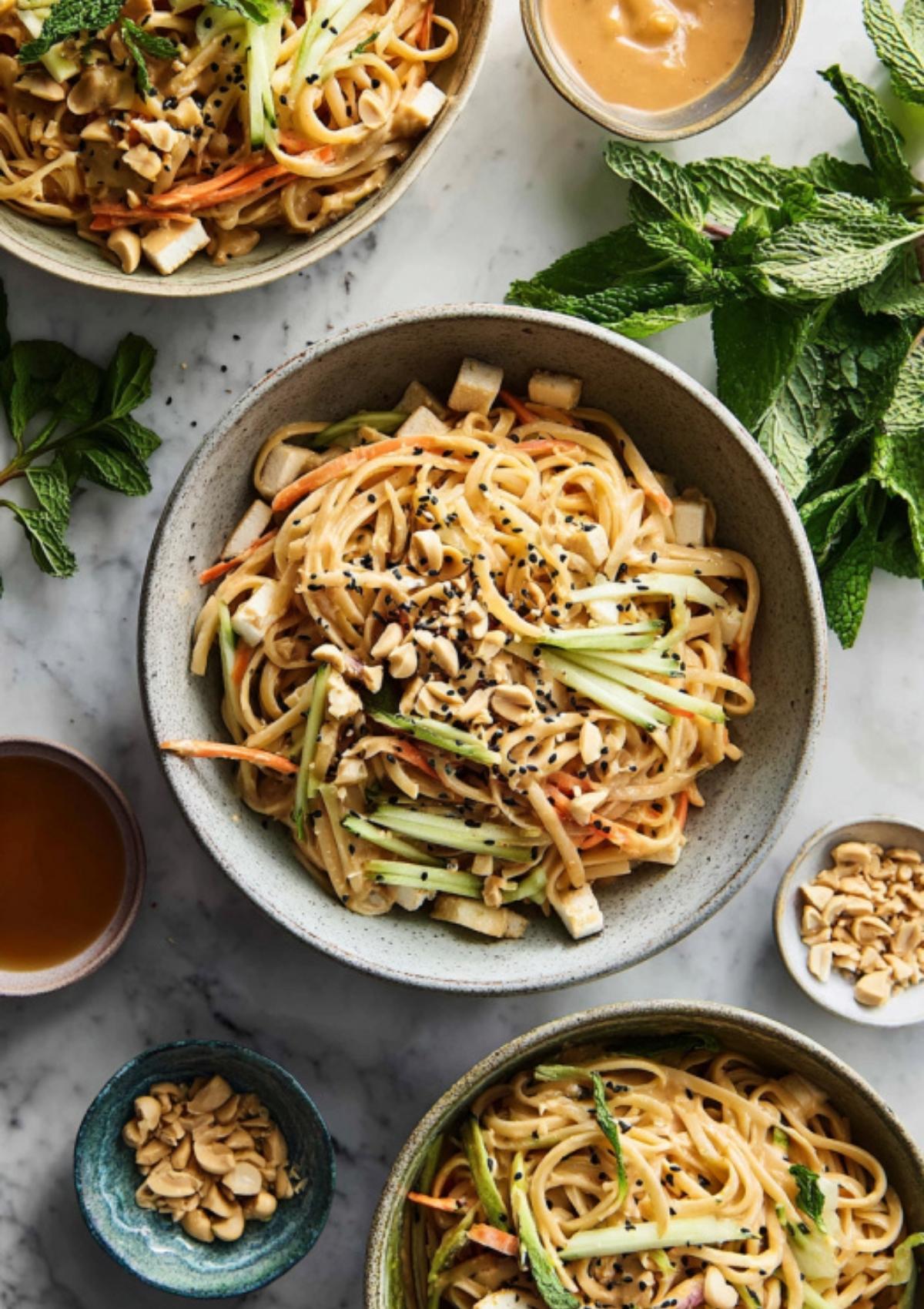 Bowls of Thai peanut noodles topped with cucumber, carrots, tofu, black sesame seeds, and crushed peanuts.