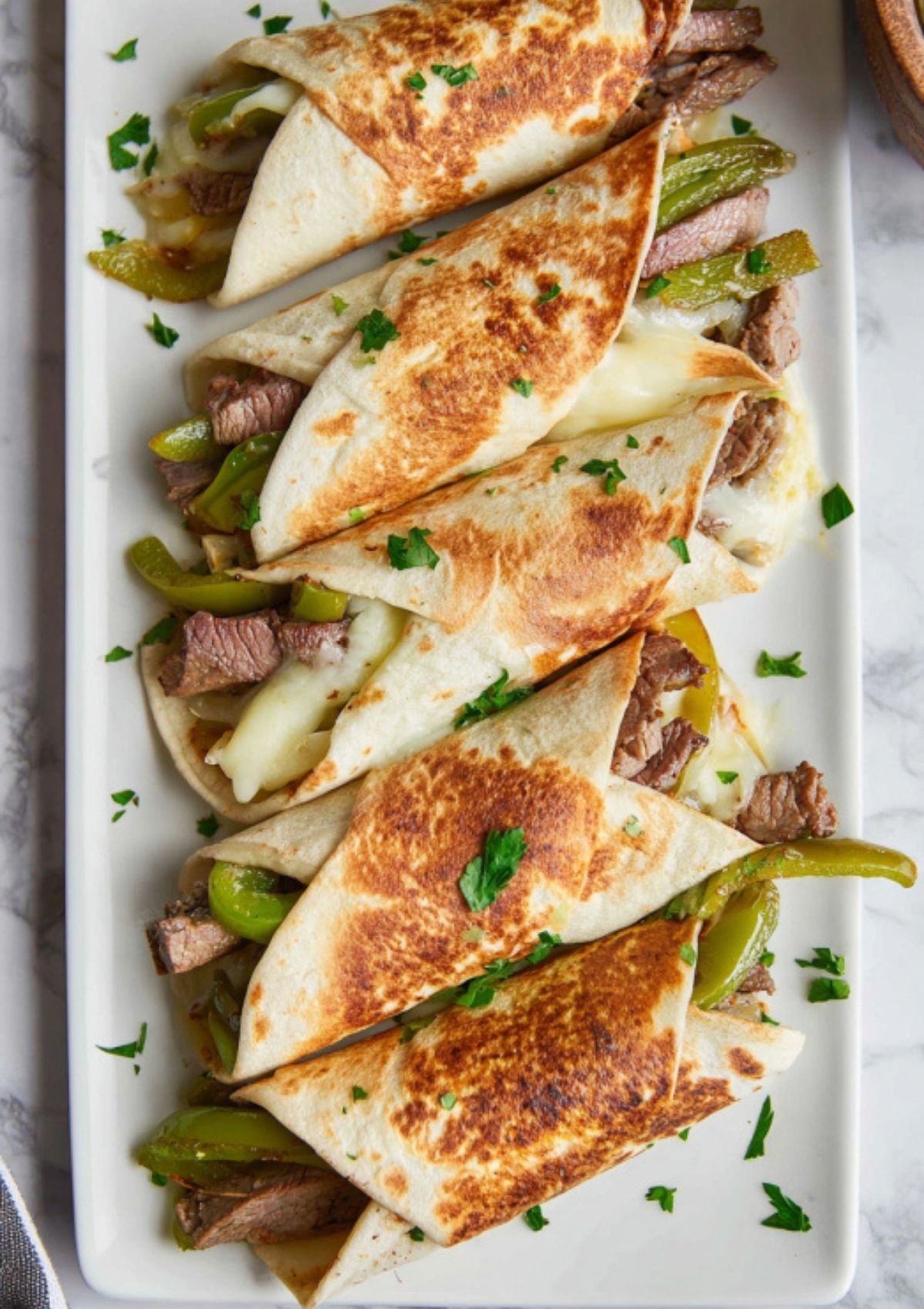 Platter of toasted Philly cheesesteak wraps, filled with steak, green peppers, and melted cheese, garnished with fresh parsley.