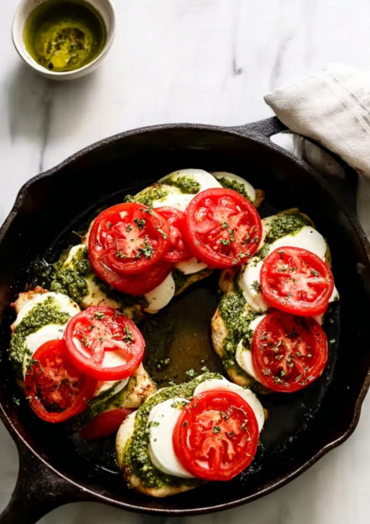 Chicken breasts in a skillet layered with pesto, mozzarella, and fresh tomato slices, ready to bake.