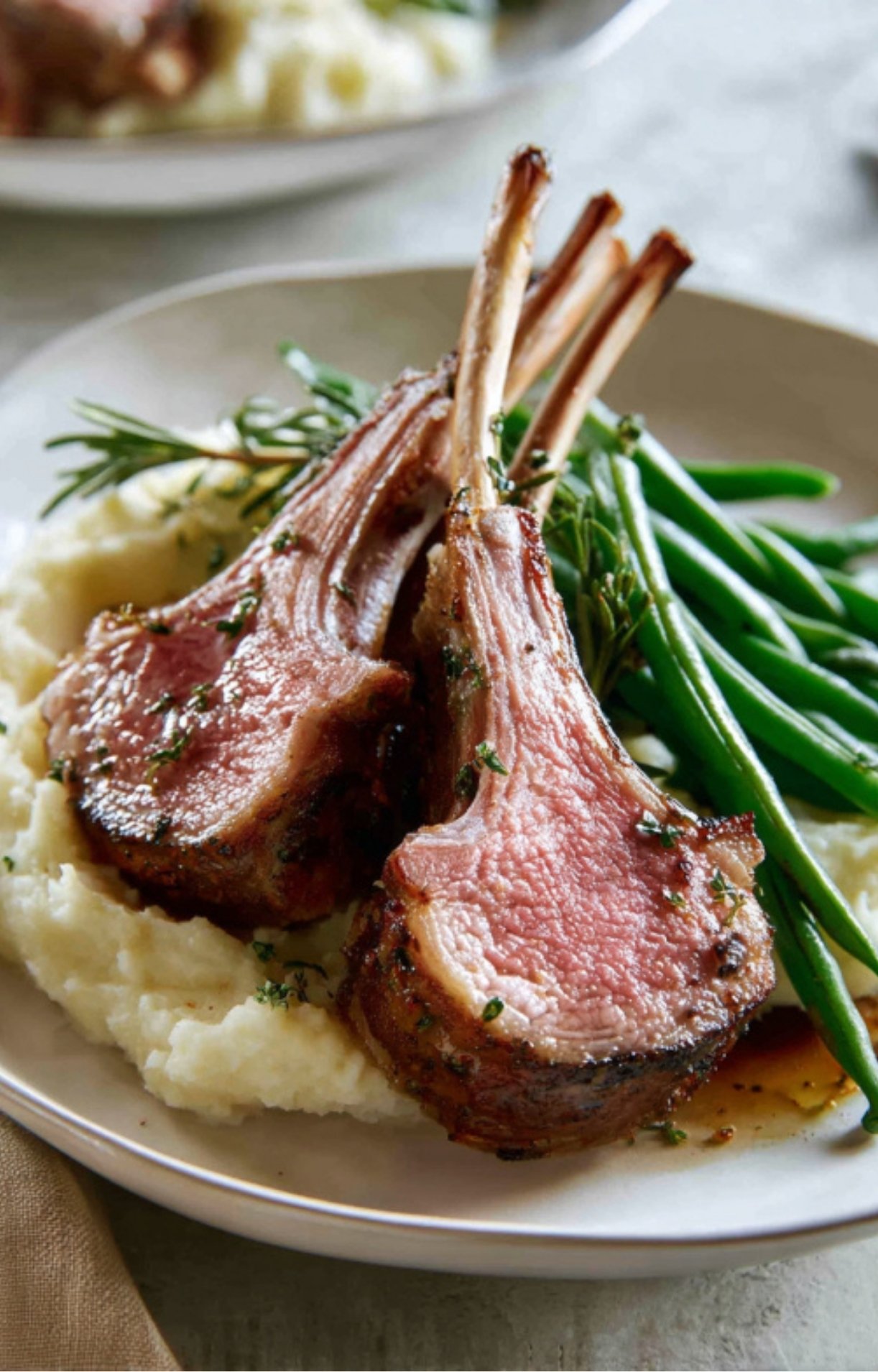 Slices of roasted rack of lamb served over creamy mashed potatoes and green beans on a dinner plate.