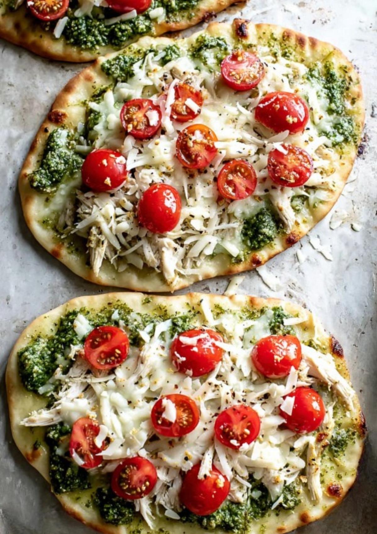 Flatbreads topped with pesto, shredded chicken, mozzarella, and cherry tomatoes on a baking sheet, ready for the oven.