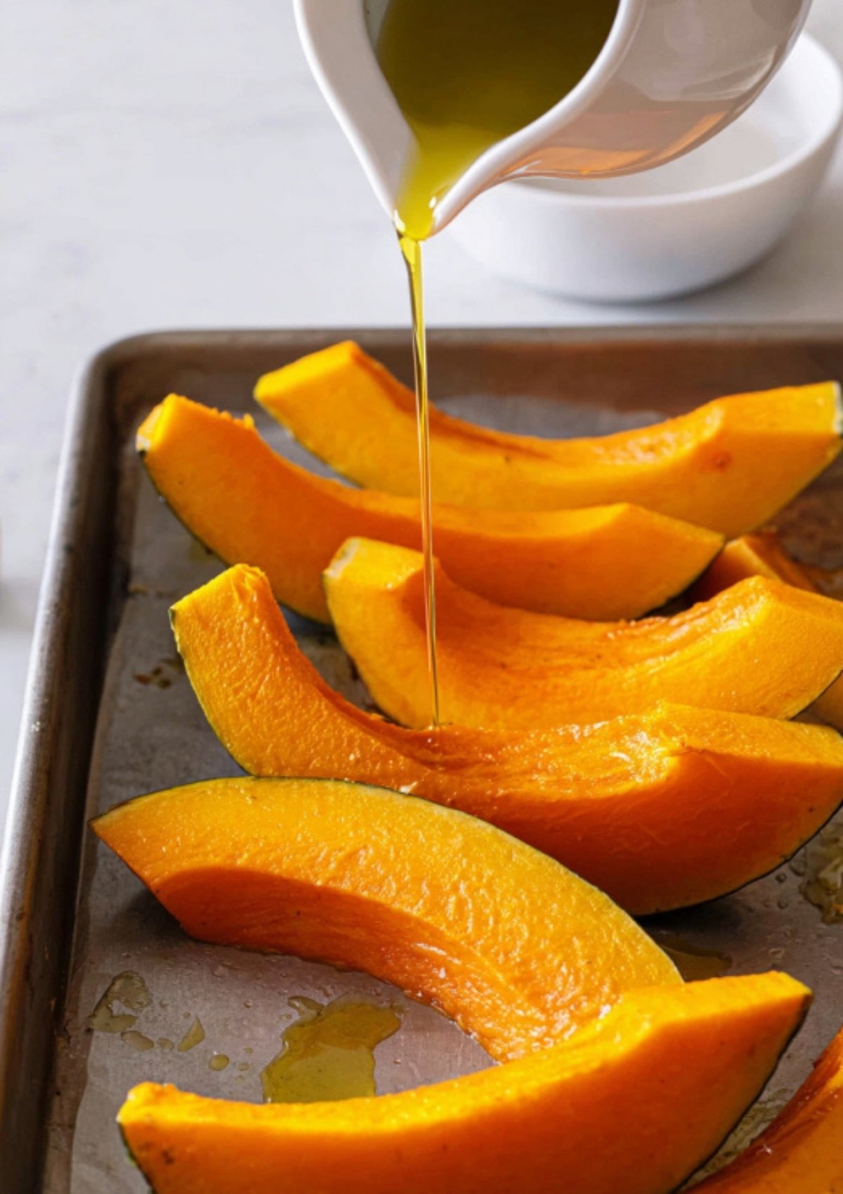Raw pumpkin wedges on a baking sheet, being drizzled with golden olive oil, ready for the oven.
