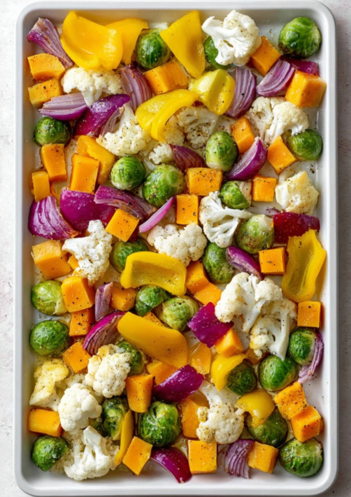 Overhead view of colorful chopped vegetables—brussels sprouts, red onion, yellow bell pepper, cauliflower, and butternut squash—arranged on a baking sheet, ready for roasting.