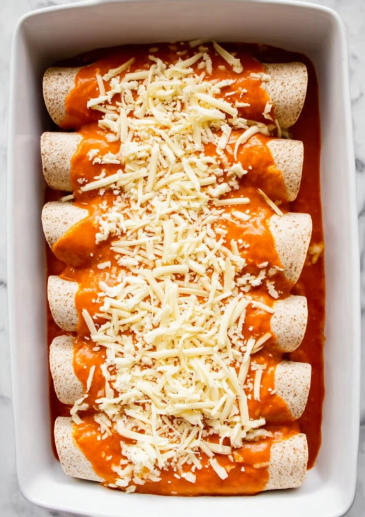 Unbaked black bean enchiladas in a white baking dish, topped with enchilada sauce and shredded cheese, ready to go in the oven.