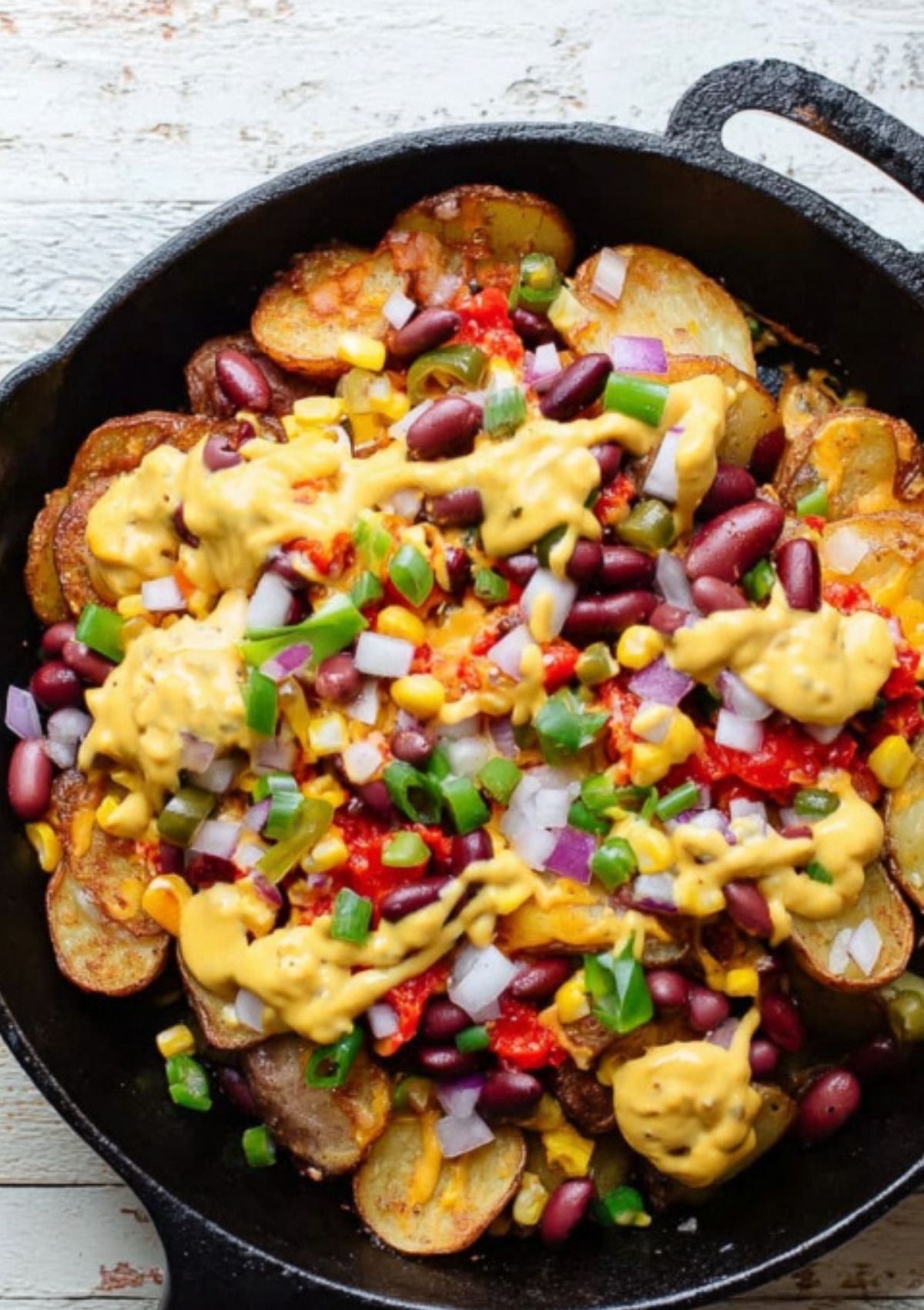 Skillet of loaded baked potato nachos covered with cheese sauce, kidney beans, salsa, corn, chopped red onions, and green onions.