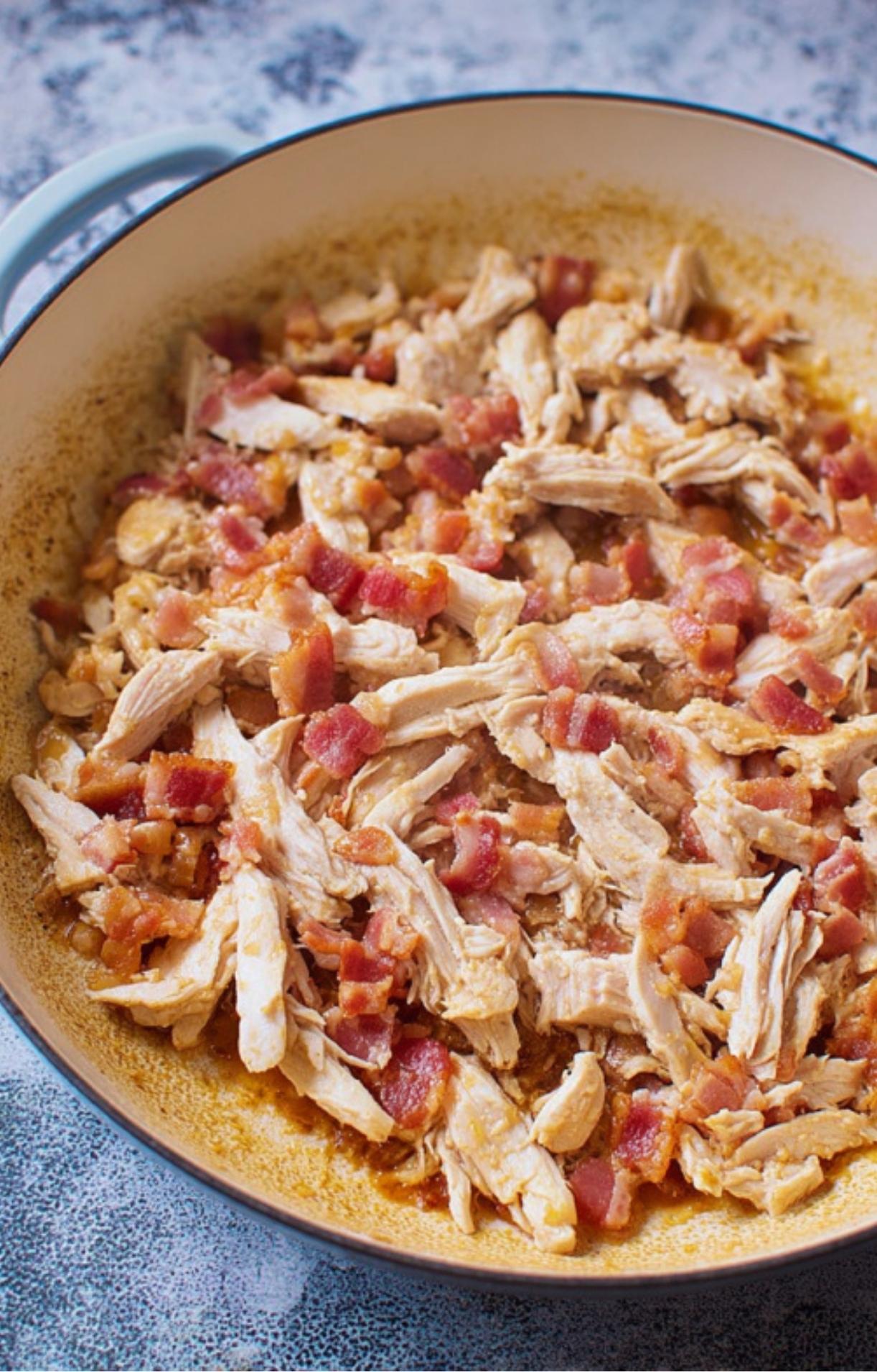 Closeup of shredded leftover roast chicken and crispy bacon pieces sizzling in a large cream-colored skillet.