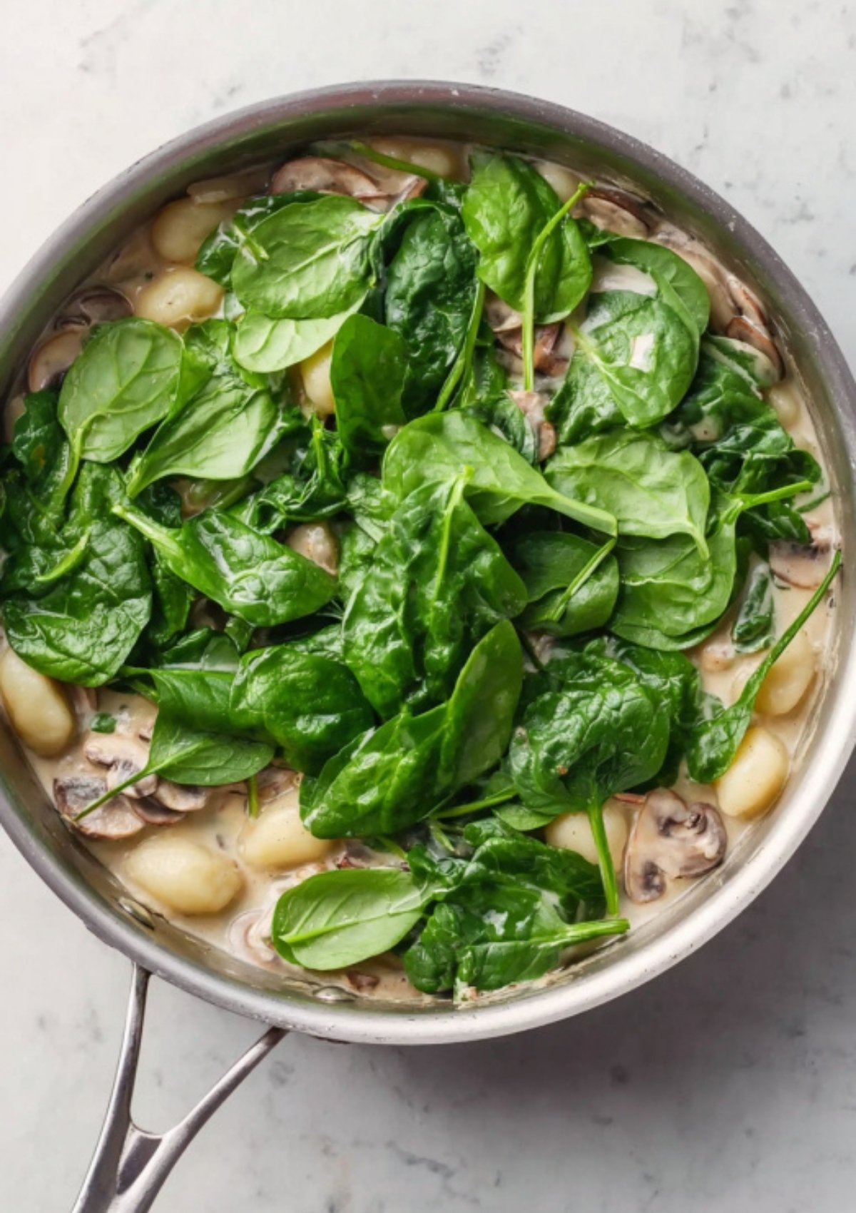 Unmixed skillet of creamy gnocchi, sautéed mushrooms, and vibrant fresh spinach leaves ready to be stirred. Simple vegetarian dinner idea.