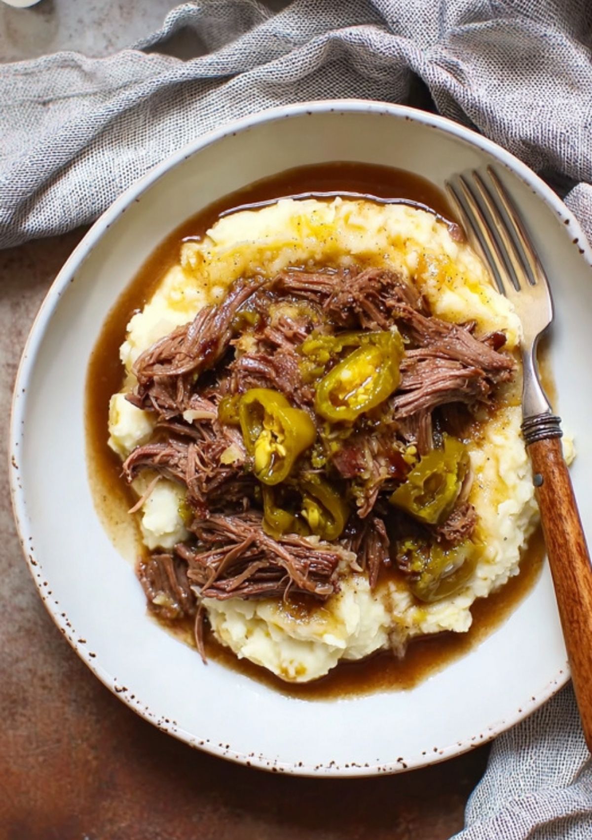 Overhead view of Mississippi pot roast served on creamy mashed potatoes with brown gravy and sliced pepperoncini, styled on a rustic plate with a fork.