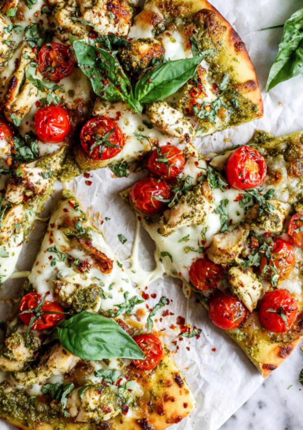 Sliced pesto chicken flatbread with fresh basil and cherry tomatoes on parchment paper.