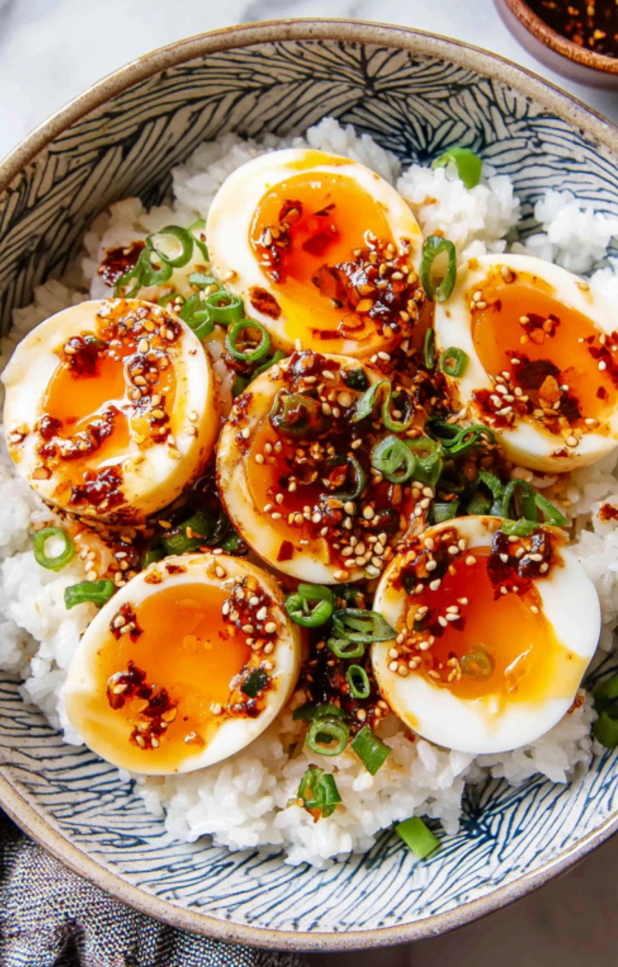 Bowl of steamed white rice topped with halved Korean mayak eggs (soy sauce marinated eggs) with jammy yolks, drizzled with chili oil, sprinkled with sesame seeds and sliced green onions.