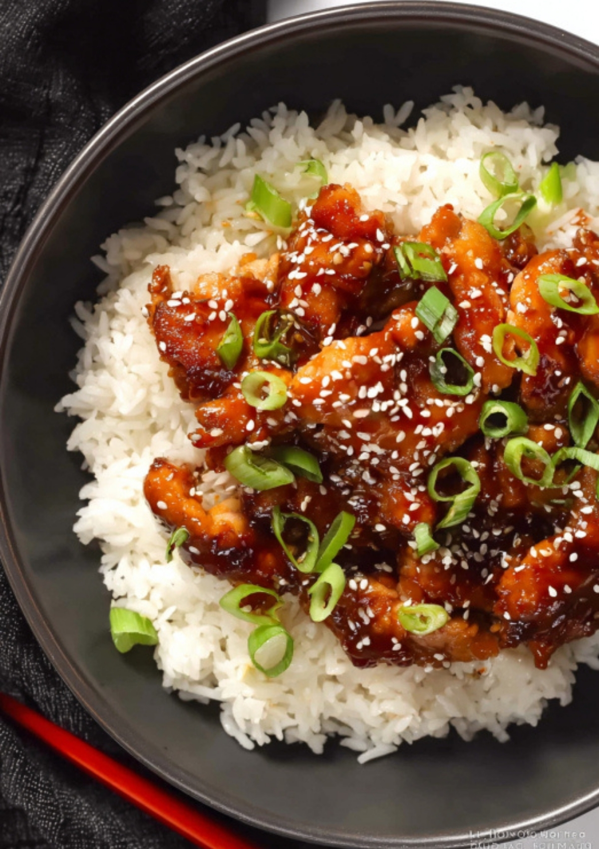 A rice bowl topped with sticky soy glazed chicken, green onion slices, and sesame seeds, ready to eat with chopsticks.