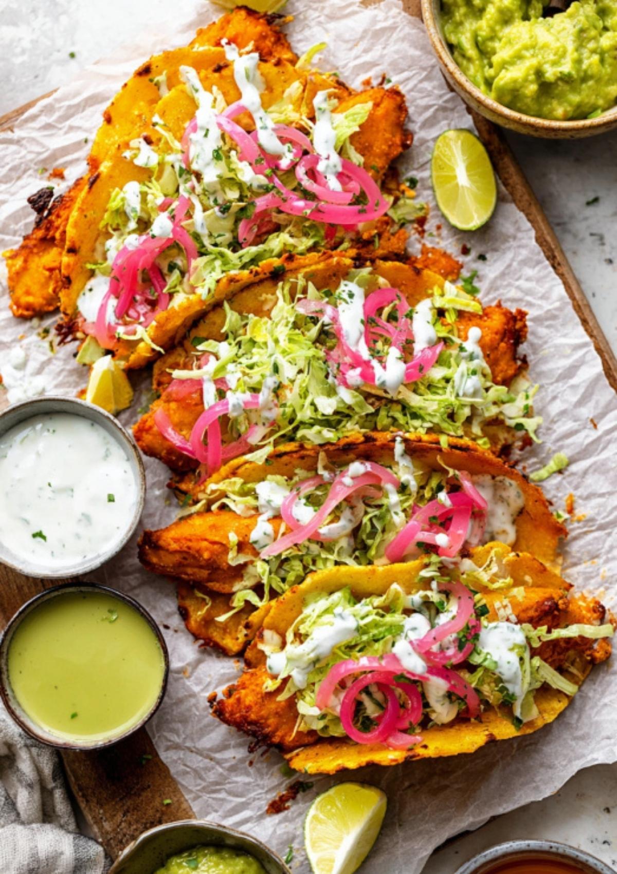 Crispy buffalo chicken tacos on parchment paper served with sauces, lime wedges, and guacamole.