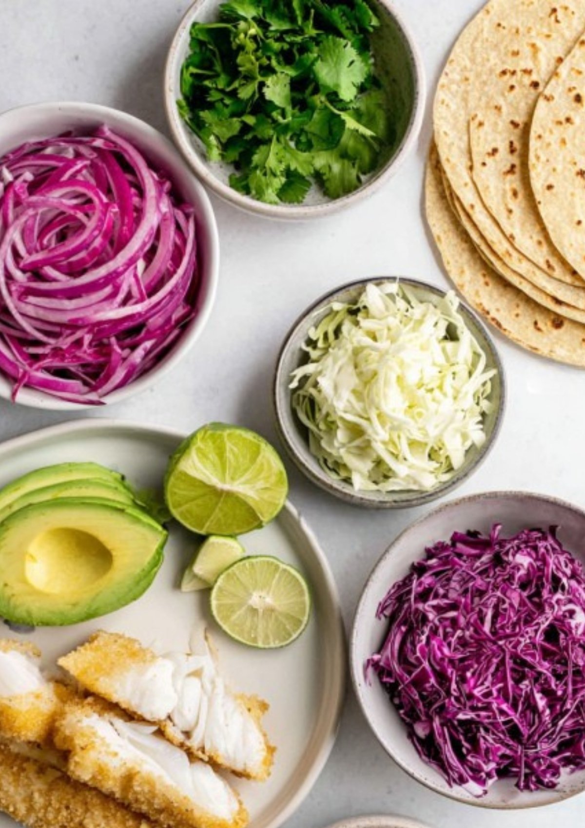 Overhead shot of ingredients for crispy fish tacos, including sliced avocado, lime, shredded red and green cabbage, red onion, cilantro, corn tortillas, and flaky breaded white fish.
