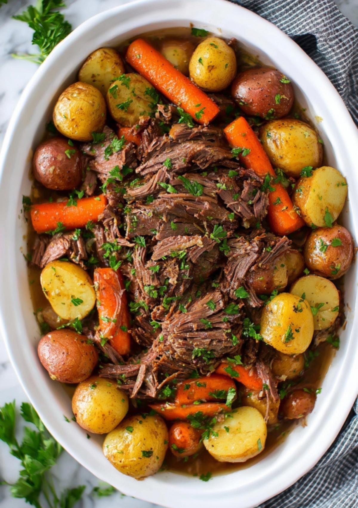 Shredded pot roast served with carrots and baby potatoes in a white serving dish, garnished with parsley.