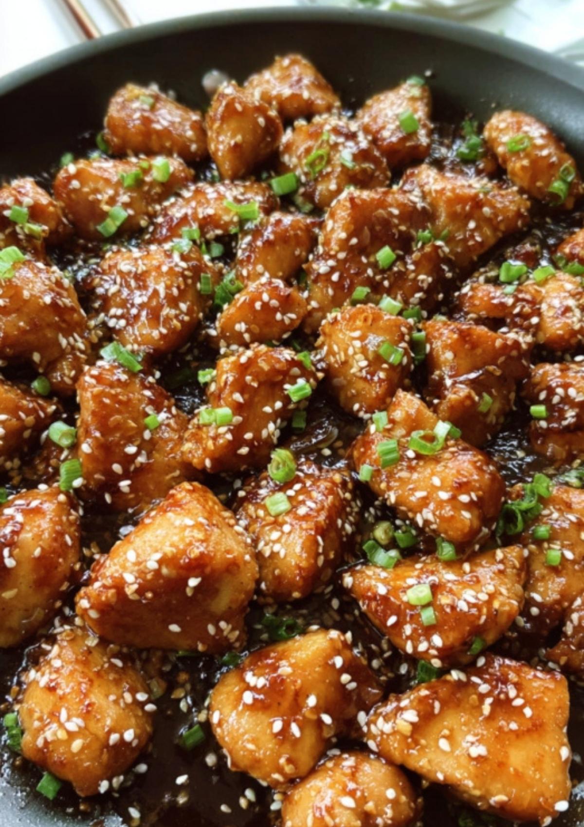 Bite-sized chicken pieces coated in sesame sauce and garnished with sesame seeds and scallions in a skillet.
