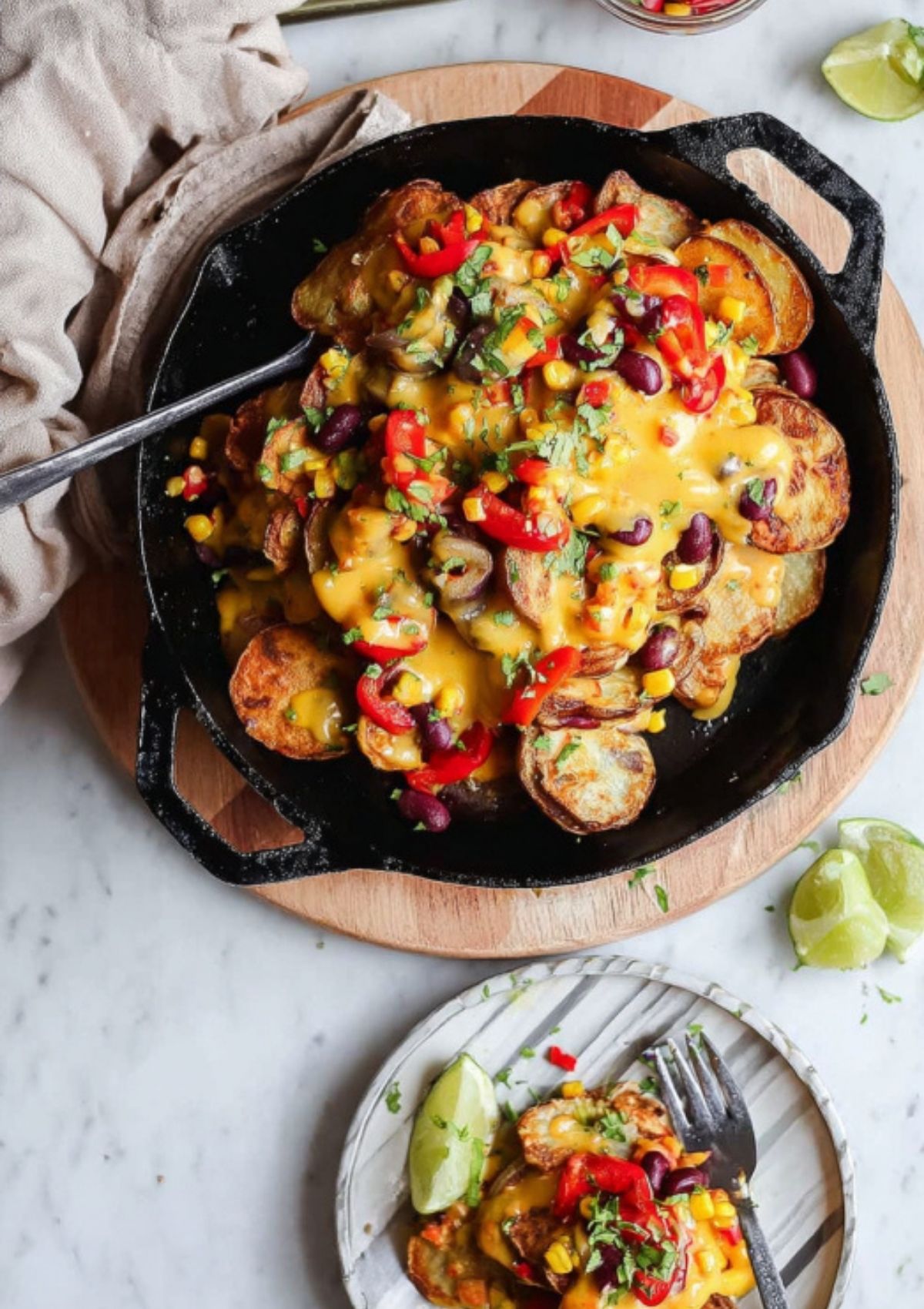 Overhead shot of a cast iron skillet filled with loaded baked potato nachos, topped with cheddar cheese, beans, corn, red peppers, and fresh cilantro, served with lime wedges.
