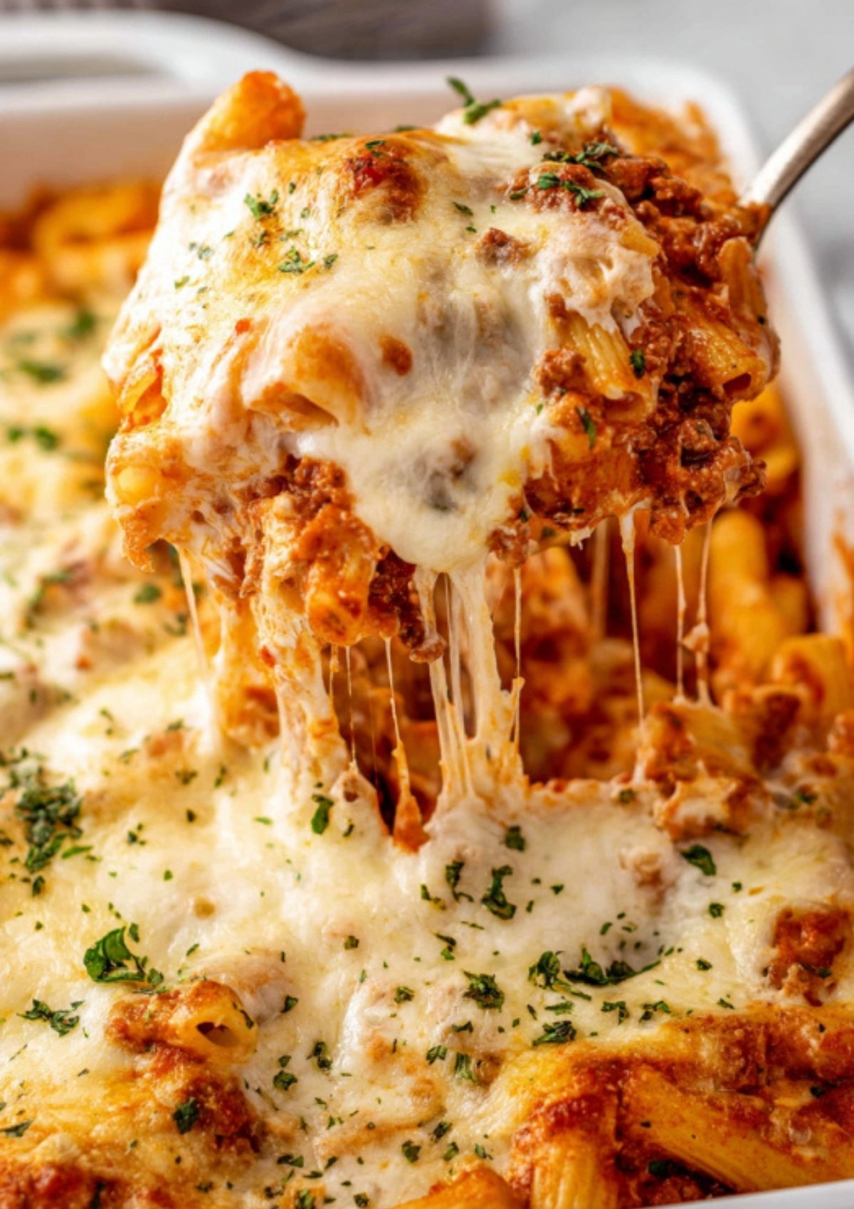 Cheesy baked ziti with ricotta being lifted from a casserole dish topped with melted mozzarella and parsley.