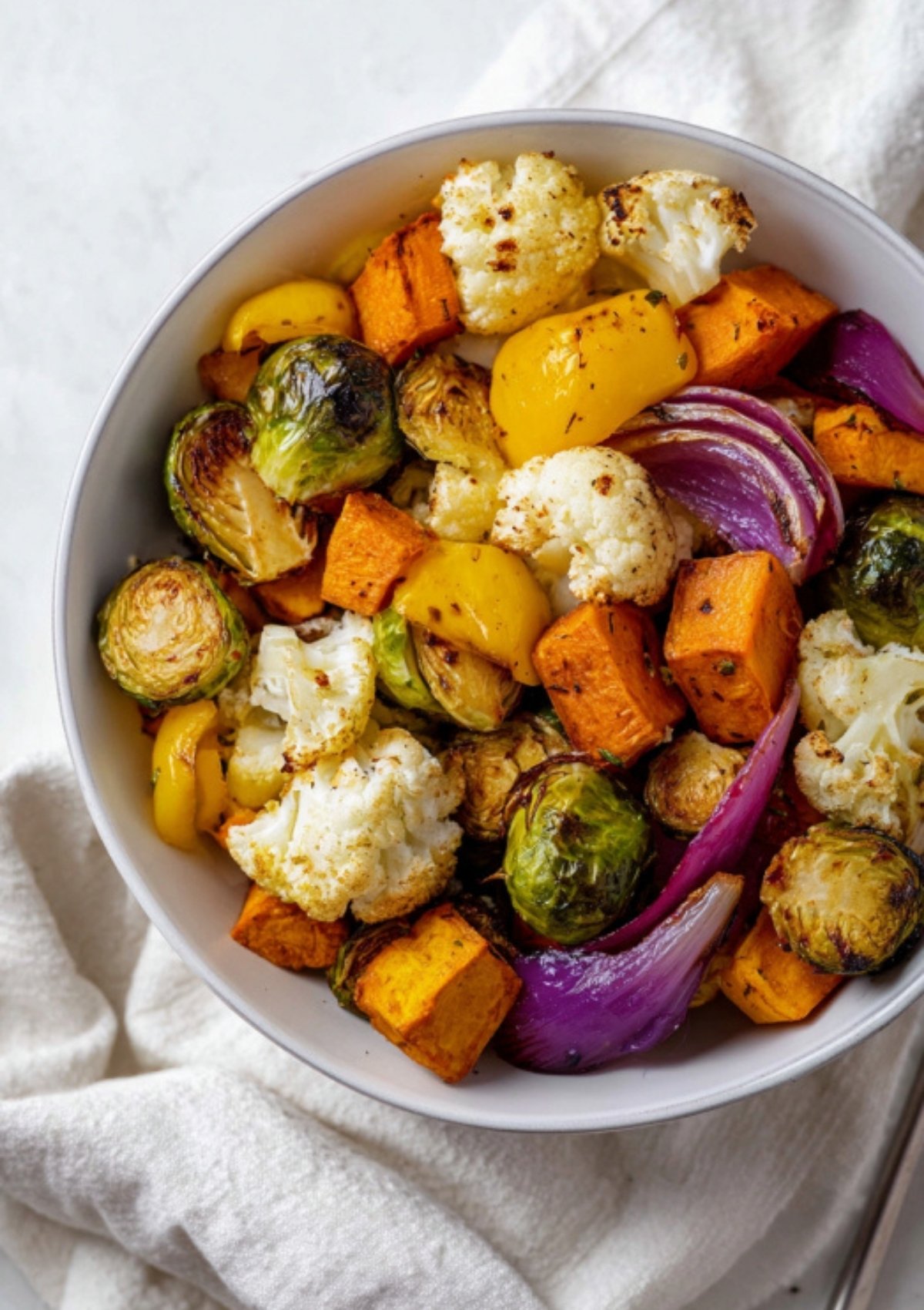Closeup of a white bowl filled with oven-roasted red onion, cauliflower, yellow bell pepper, butternut squash, and brussels sprouts.