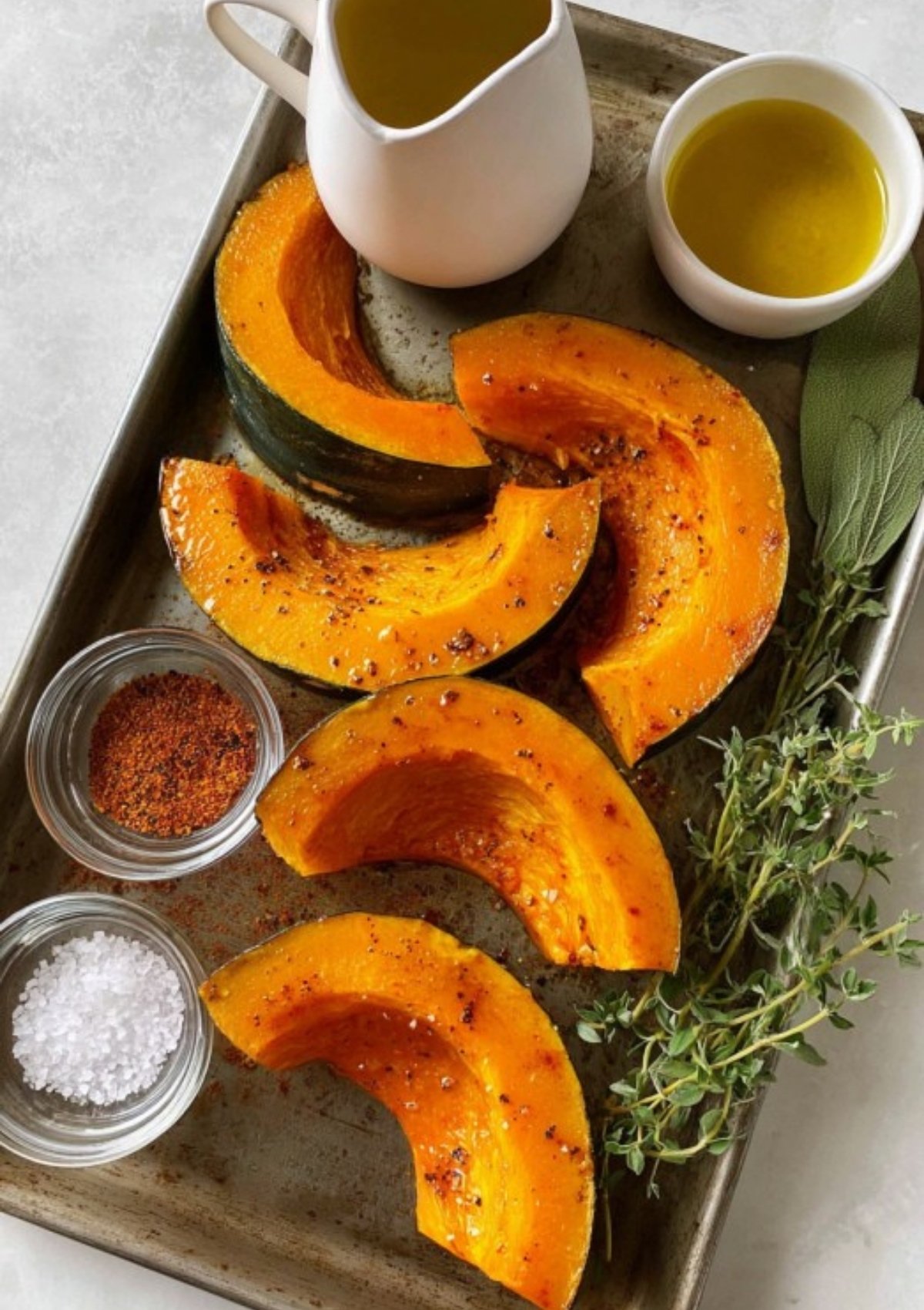 Raw pumpkin wedges on a baking sheet, ready to roast, surrounded by olive oil, fresh herbs, sea salt, and spices for a flavorful fall side dish.