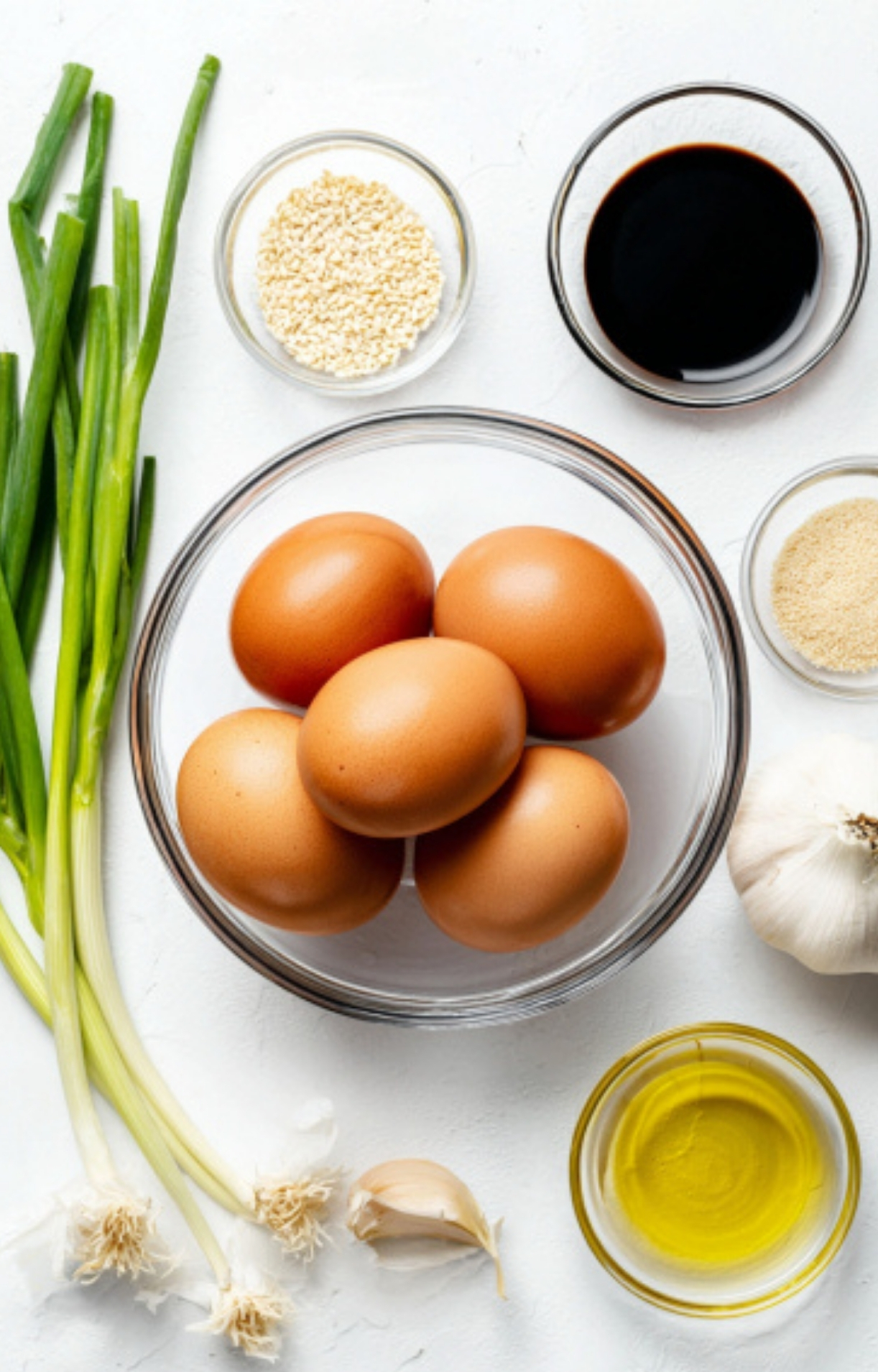 Flat lay of ingredients for Korean egg marinade: fresh eggs, green onions, sesame seeds, soy sauce, garlic, sugar, and oil on a white background.