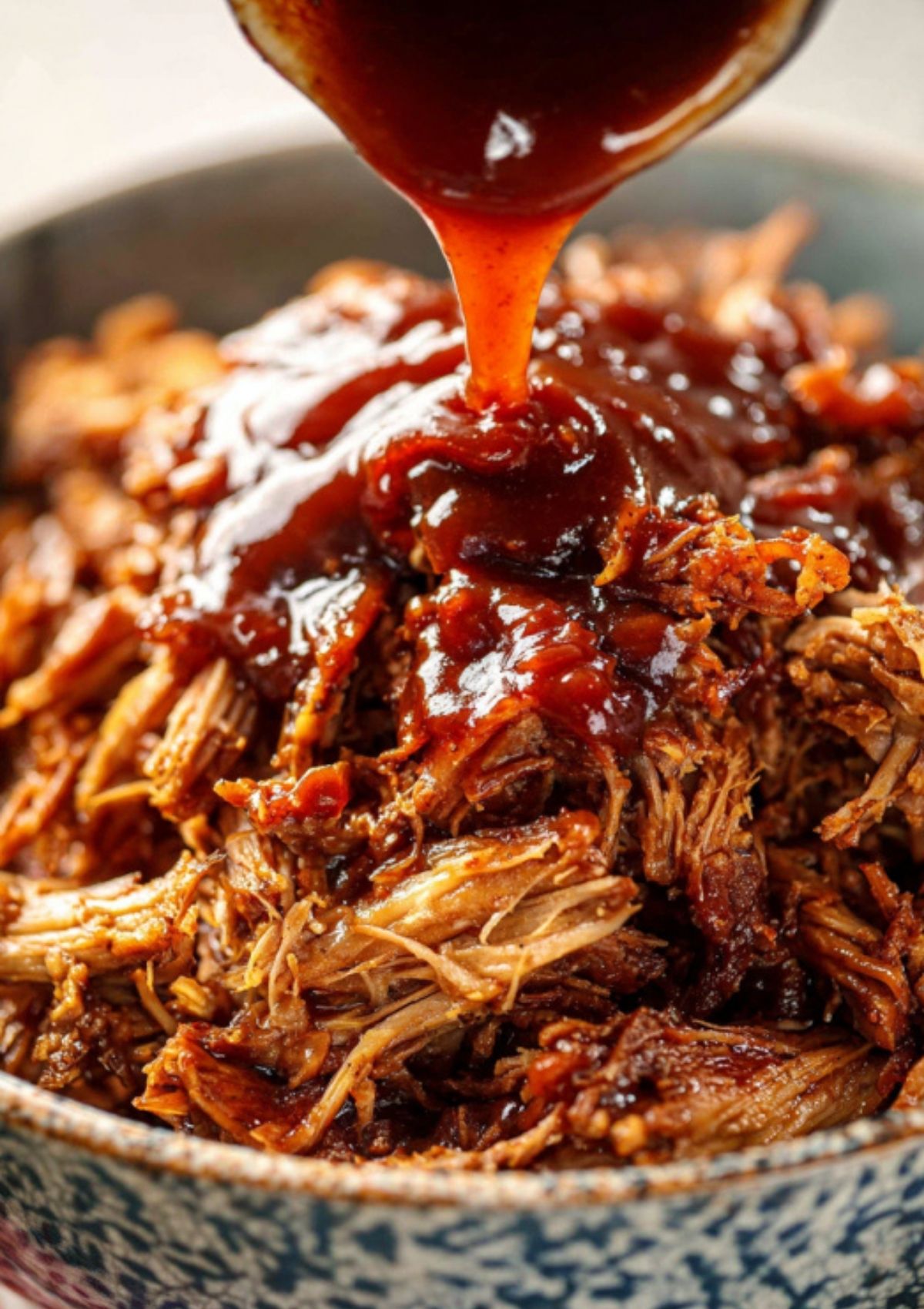 Thick barbecue sauce being poured over shredded pork in a serving bowl.