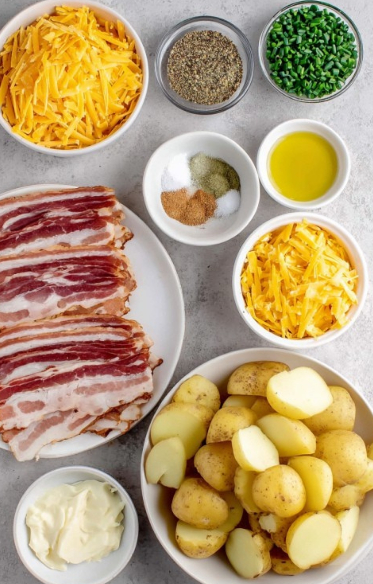 Bowls of shredded cheddar, sliced bacon, chopped potatoes, chives, spices, and olive oil prepped for making cheesy bacon ranch potatoes.